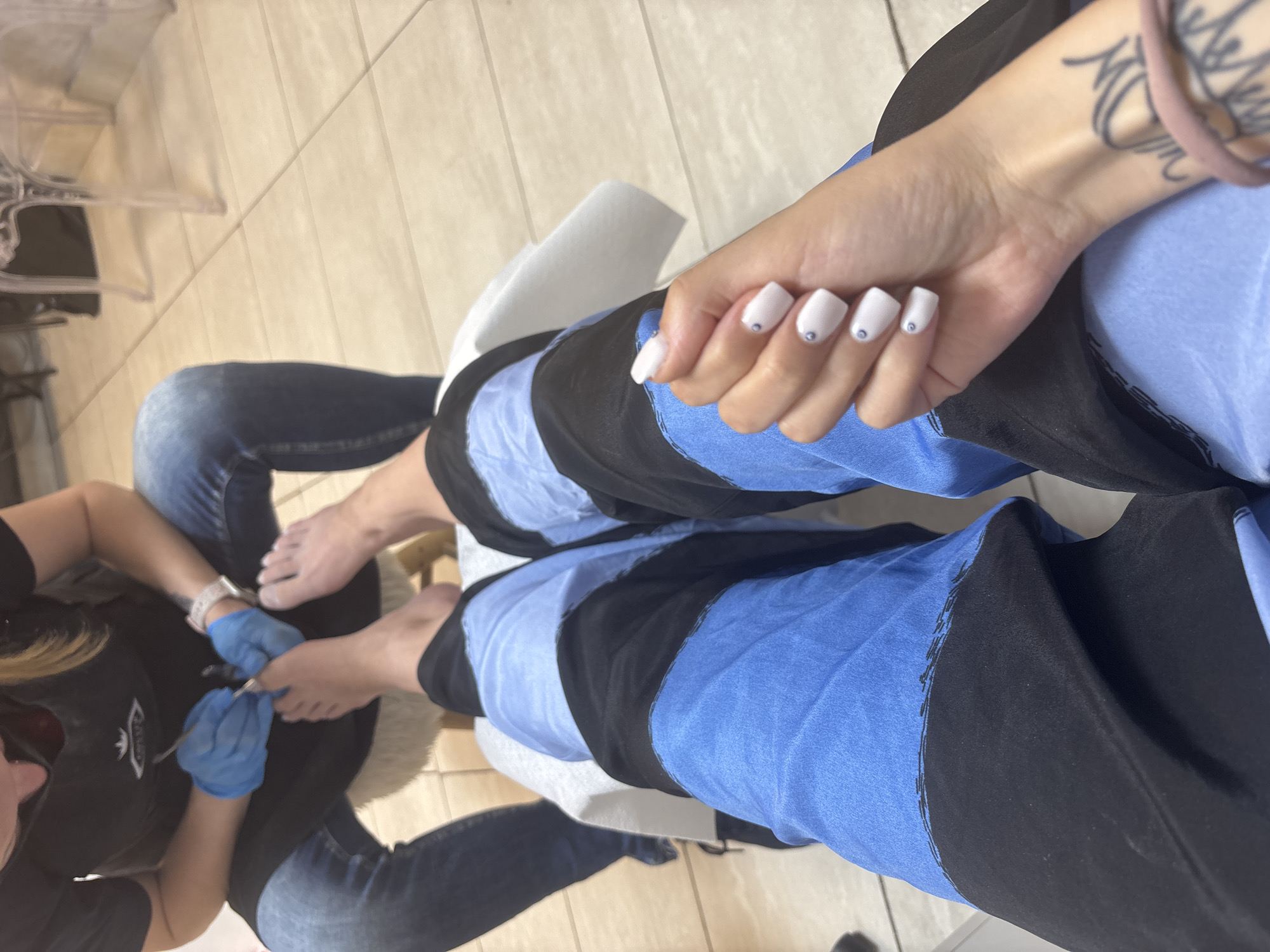 A person is receiving a pedicure. The person is wearing blue and black pants. A nail technician in a black t-shirt and blue gloves is performing the pedicure. The fingernails of the person receiving the pedicure are white with small gem-like embellishments. The location is unknown and there is no way to tell if the scene is in Sofia, Bulgaria, or any other city.