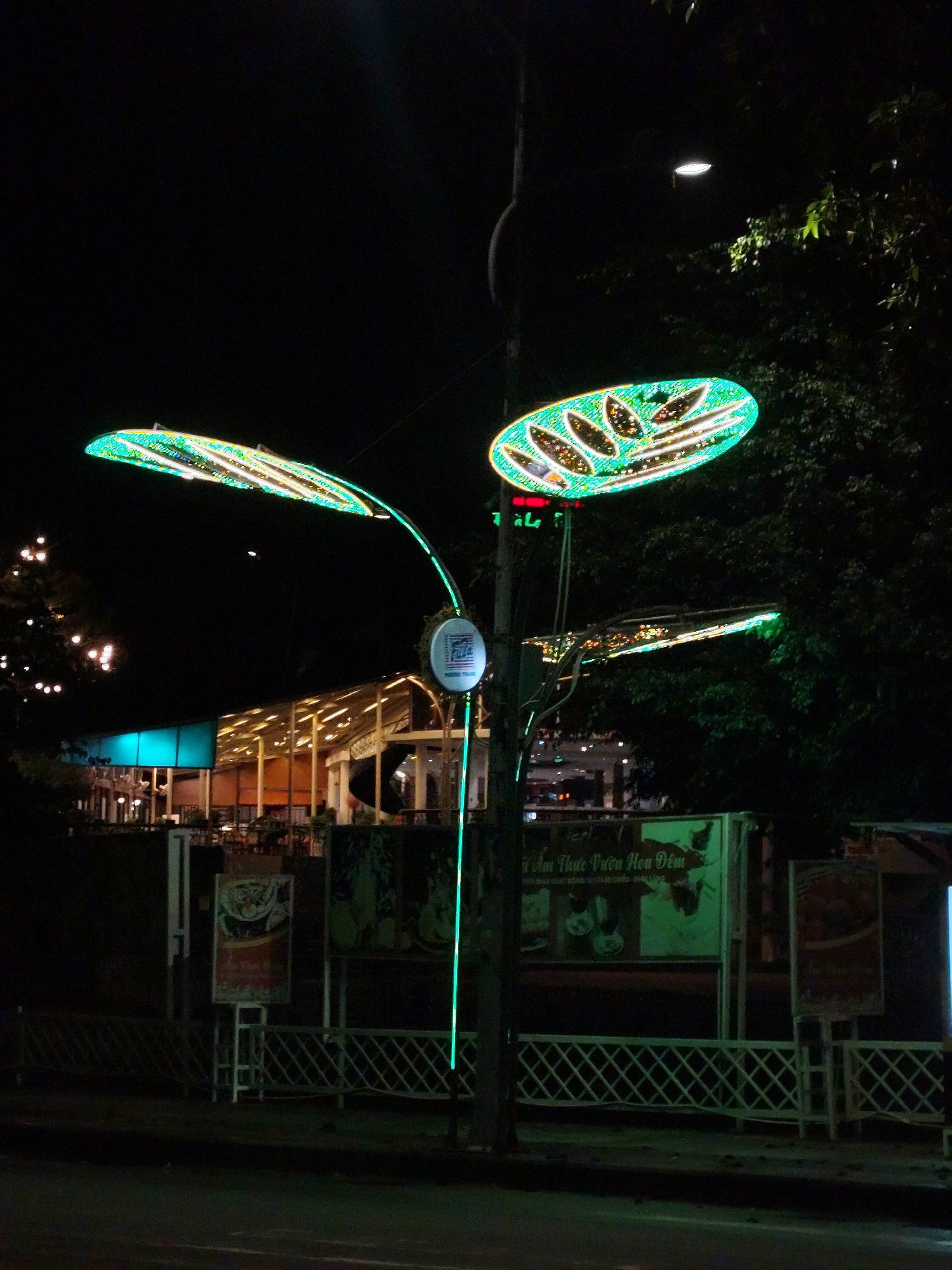 At night in Da Lat, Vietnam, a decorative streetlamp resembling illuminated leaves stands near the sidewalk. Behind it, a restaurant with a brightly lit, open-air structure can be seen. A metal fence runs along the sidewalk, and several advertisement boards are visible behind it. The street is dark and wet.