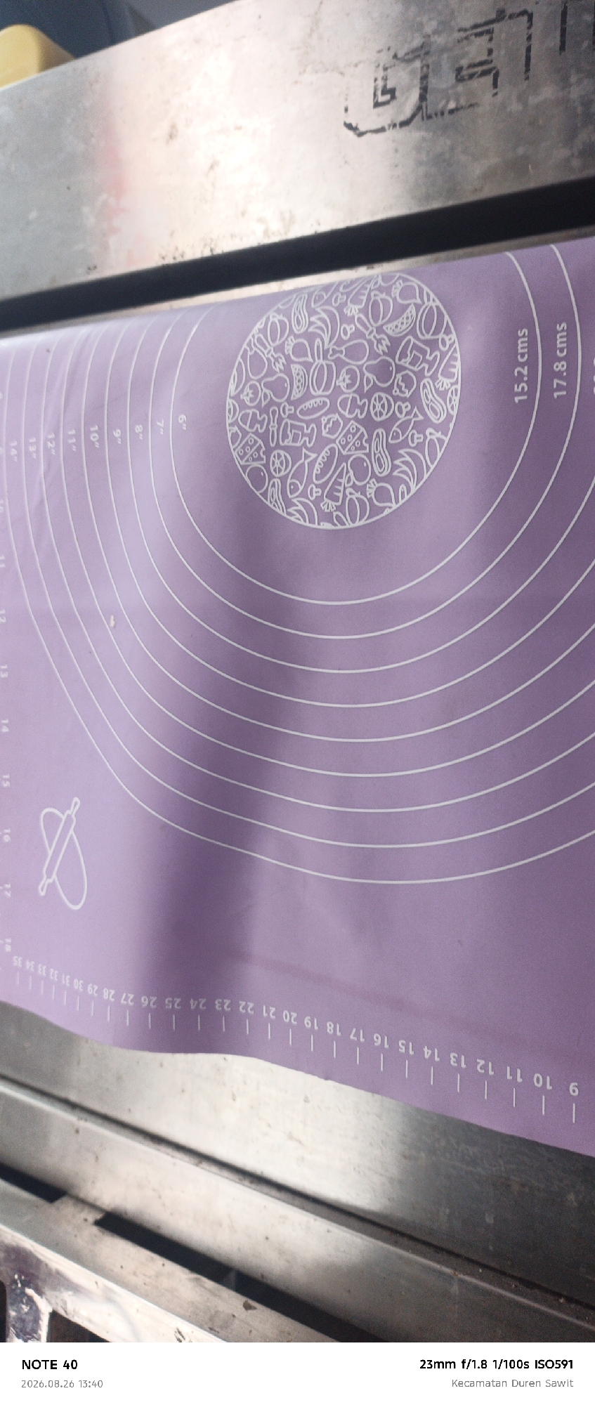 The image shows a close-up view of a purple baking mat with white markings. The mat has concentric circles and radial lines, indicating measurements in inches and centimeters. There is a circular pattern of food illustrations at the center of the mat. The mat is placed on a metallic surface, likely a countertop or table. The upper portion of the image shows a portion of a metallic object with some markings. The bottom of the image contains text overlay information, including "NOTE 40", "2026.08.26 13:40", "23mm f/1.8 1/100s ISO591", and "Kecamatan Duren Sawit". This suggests the photo was taken in the Duren Sawit District, East Jakarta, Indonesia.