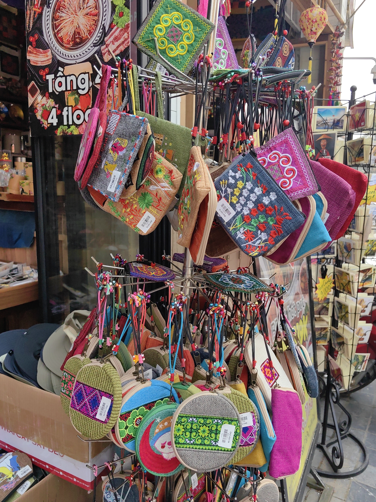 The image depicts a close-up view of a street-side merchandise display in Sa Pa, Vietnam. The central subject is a multi-tiered metal display rack exhibiting numerous small, handcrafted textile items, primarily pouches or coin purses. These items feature diverse shapes (rectangular, circular, semi-circular), vibrant colors, and intricate embroidery with floral, geometric, and abstract patterns. Many items have white price tags attached, some showing prices in Vietnamese Dong (e.g., "35,000 VND").
In the background to the left, a large promotional sign for a food establishment is visible, featuring images of hotpot ingredients (meat, mushrooms) and text including "Tầng 4 floo" (indicating "4th Floor"). Further left, reflections in a window show glimpses of an indoor shop interior with shelves and additional merchandise.
To the right, another black metal display rack holds numerous postcards, some depicting landscapes and a person wearing a traditional Vietnamese conical hat. A sign near these postcards reads "Postcards 9.000 VND". Above, a decorative yellow lantern with a floral pattern hangs from an awning or structure. The environment suggests an active commercial area, likely a market or shop entrance, on a street in Sa Pa. No individuals are visible.