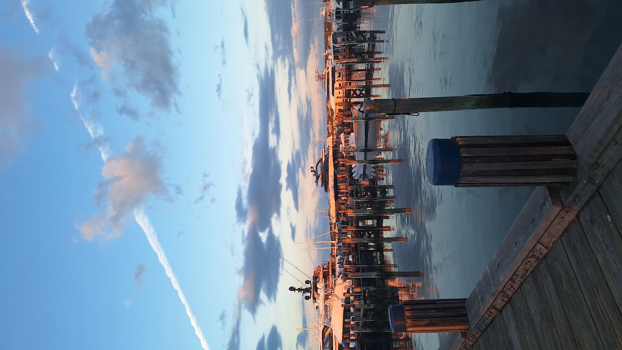The photograph captures a serene harbor scene at dusk in Nantucket, Massachusetts. Several luxury yachts are docked along a wooden pier, their sleek forms illuminated by the warm, golden light of the setting sun. The sky above is a canvas of soft blues and pinks, with wispy clouds catching the last rays of daylight and a distinct contrail streaking across the upper left quadrant. The water in the harbor is calm, reflecting the boats, pier, and the colorful sky. Wooden pilings, some capped with blue, line the edge of the dock, and the wooden planks of the pier are visible in the foreground. The overall atmosphere is one of tranquility and coastal elegance.