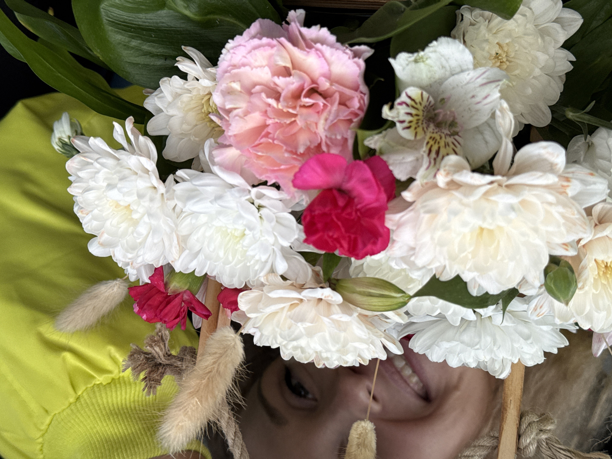 A close-up, upside-down shot reveals a person wearing a yellow shirt, almost entirely obscured by a floral headdress. The headdress is composed of various flowers including white chrysanthemums, a pink carnation, and a few smaller red flowers. Green leaves are visible among the flowers. The lower portion of the person's face is visible beneath the floral arrangement, showing an eye, a portion of their nose, and a smiling mouth. Based solely on the visible information, the location in Perm, Russia cannot be determined.