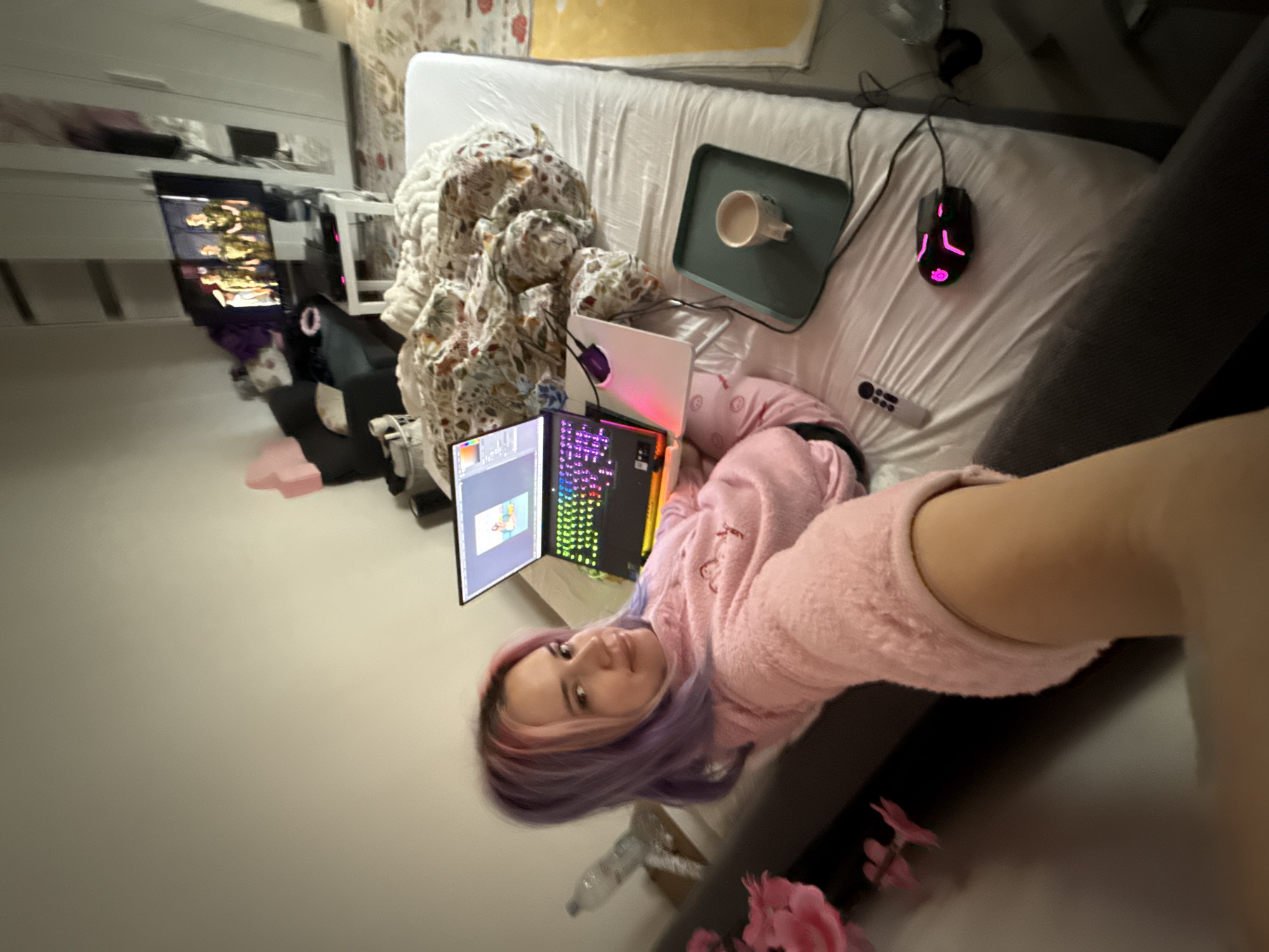 A young adult female with multi-colored (purple and pink) hair is depicted in a residential interior, likely a bedroom, within the City of Zagreb, Croatia. She is lying on a bed covered with a white fitted sheet, wearing a pink fluffy long-sleeved top and matching pajama pants. Her left arm, obscured by the camera's perspective, extends forward as she takes a selfie, looking directly at the viewer with a slight smile.
A black laptop with a brightly illuminated multi-colored (RGB) keyboard is open on a white stand next to her, displaying a digital illustration on its screen. A rumpled floral-patterned duvet or blanket rests on the bed beside the laptop. To the laptop's right, a light green rectangular tray holds a white mug containing a light-colored liquid. A white television remote control and a black gaming mouse with pink LED lighting are also present on the bed sheets. A clear plastic water bottle is visible on a small table or stand to her left.
In the background, a flat-screen television is displaying an image of three individuals in what appears to be military attire. Below the television, a dark grey sofa or chaise lounge is visible, with items piled upon it. To the far right, a large white wardrobe with reflective surfaces stands against a light-colored wall. The floor features a patterned rug or carpet. Artificial pink flowers are visible on a dark grey surface in the immediate foreground, to the left. The room is generally well-lit.