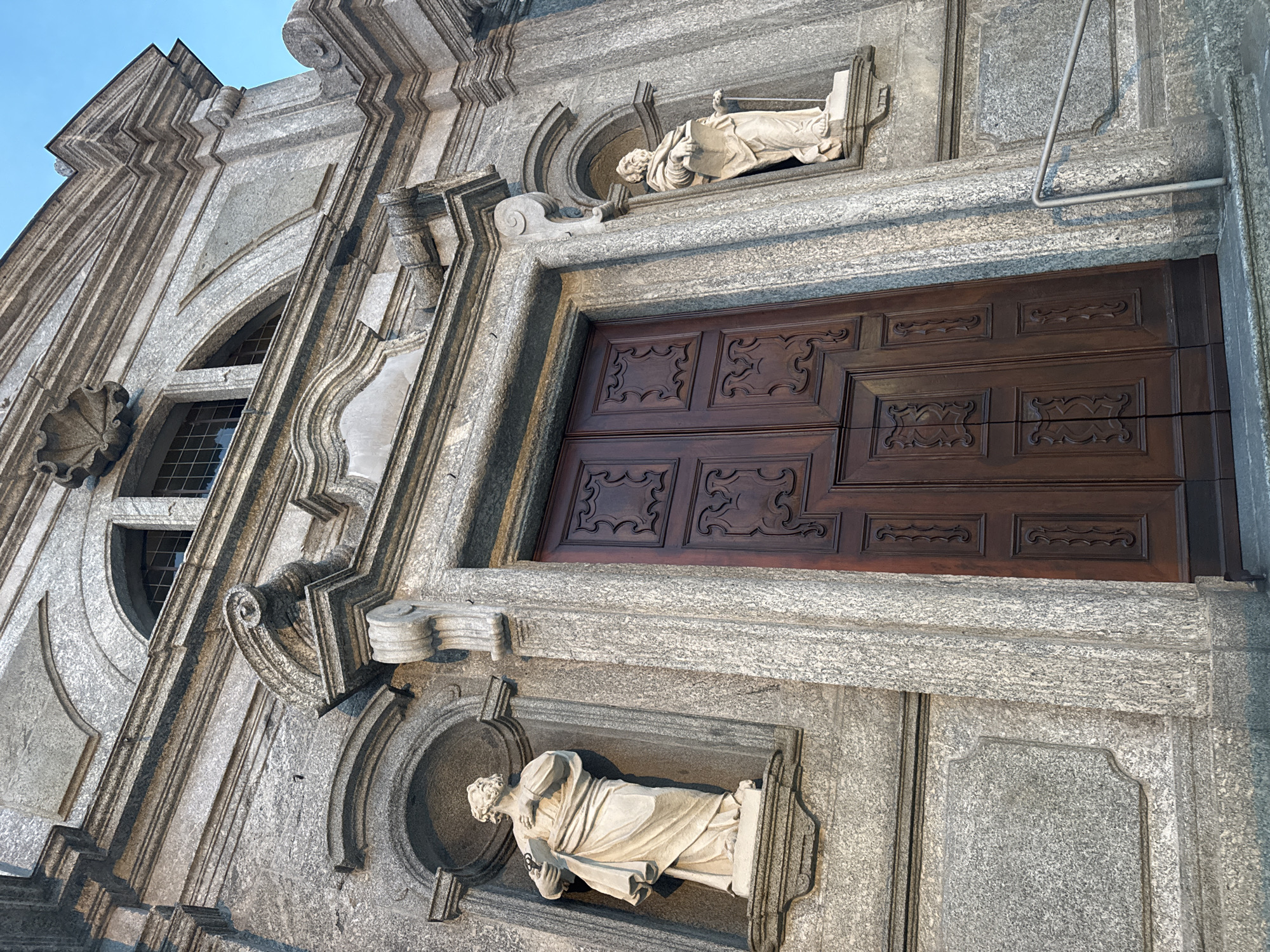 The image shows the facade of a stone building, likely a church, in Balerna, Switzerland. The building features classical architectural elements, including intricate stonework, arched windows, and statues. A large, dark wooden double door is centrally located, flanked by two niches containing white stone statues of robed figures holding books or scrolls. The stonework of the building is weathered and shows variations in color, characteristic of granite. The sky above is a clear blue. To the right of the door, a silver metal handrail leads up a set of stone steps. The perspective is low-angle, looking up at the facade.
