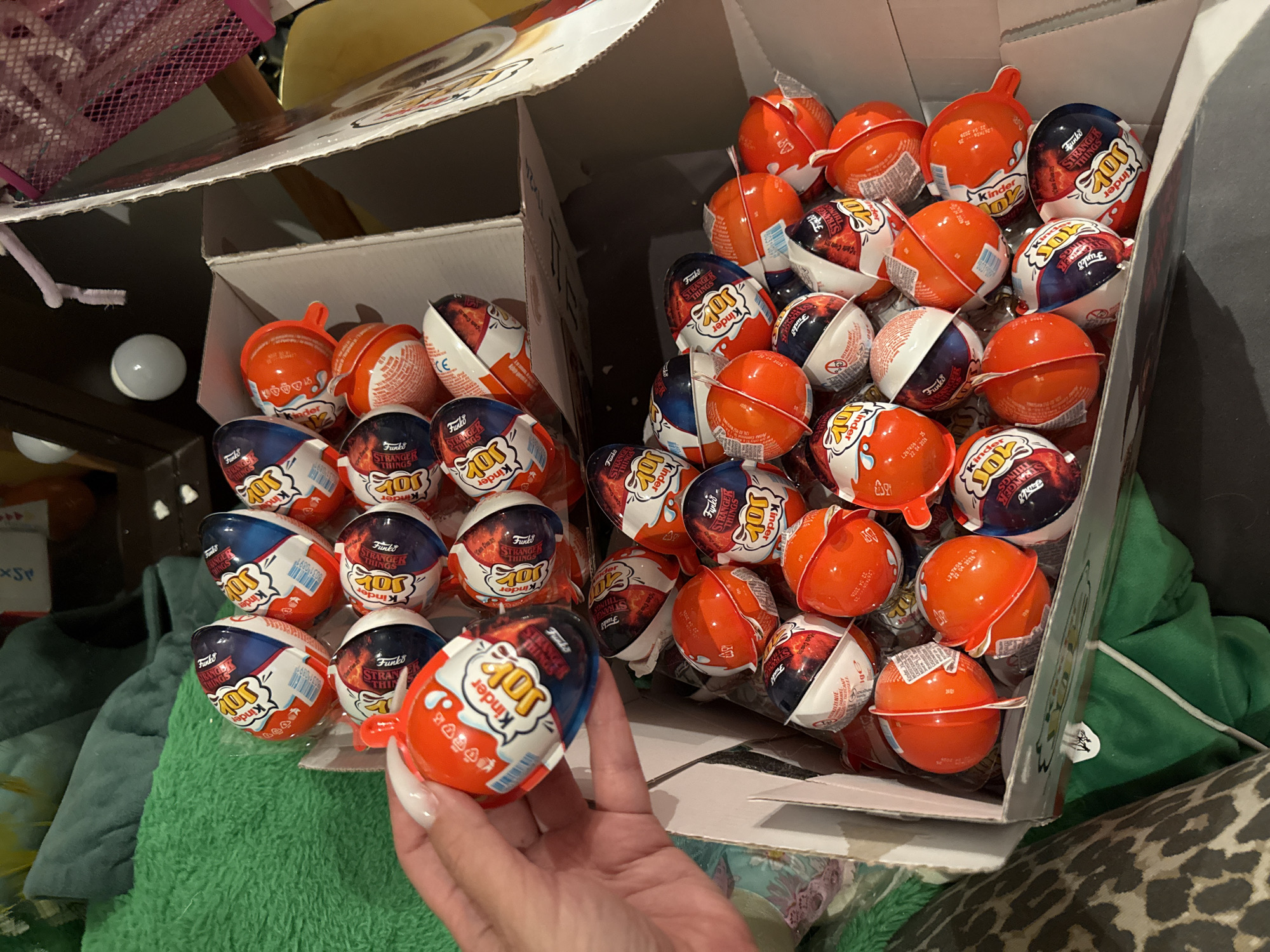 A hand with white acrylic fingernails holds an open Kinder Joy egg. The egg is orange and white with red text and a blue and red graphic. The egg is partially unwrapped, revealing the chocolate inside. The background shows a large cardboard box filled with many more Kinder Joy eggs. The eggs are individually wrapped in orange, white, blue, and red foil, many featuring graphics related to "Stranger Things." Some eggs are fully intact, while others are open, showing the chocolate inside. The box is placed on a surface, and in the background, there are blurry objects, including green fabric, a pink mesh basket, and a white spherical object. The scene appears to be an indoor setting. The location in Odesa, Ukraine, is not specified by the image content.