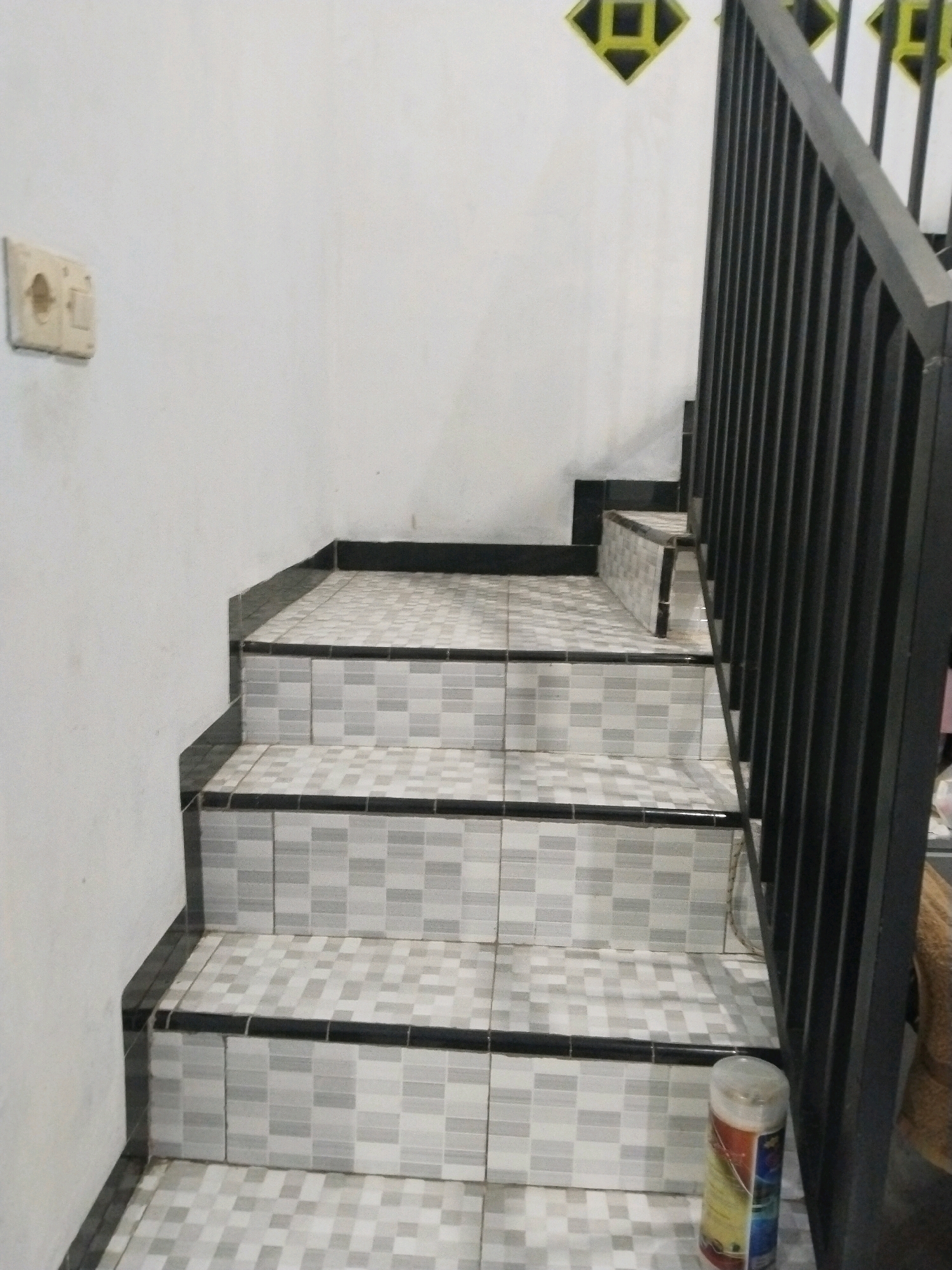 The image shows a tiled staircase with black railings leading upwards. The walls are white, and a light switch is visible on the left wall. There are decorative diamond-shaped patterns on the upper part of the wall, possibly vents. In the foreground, on the bottom step, a cylindrical object, possibly a can of aerosol, is visible. The scene appears to be in an indoor or semi-outdoor area in Tasikmadu, Indonesia, given the context provided.
