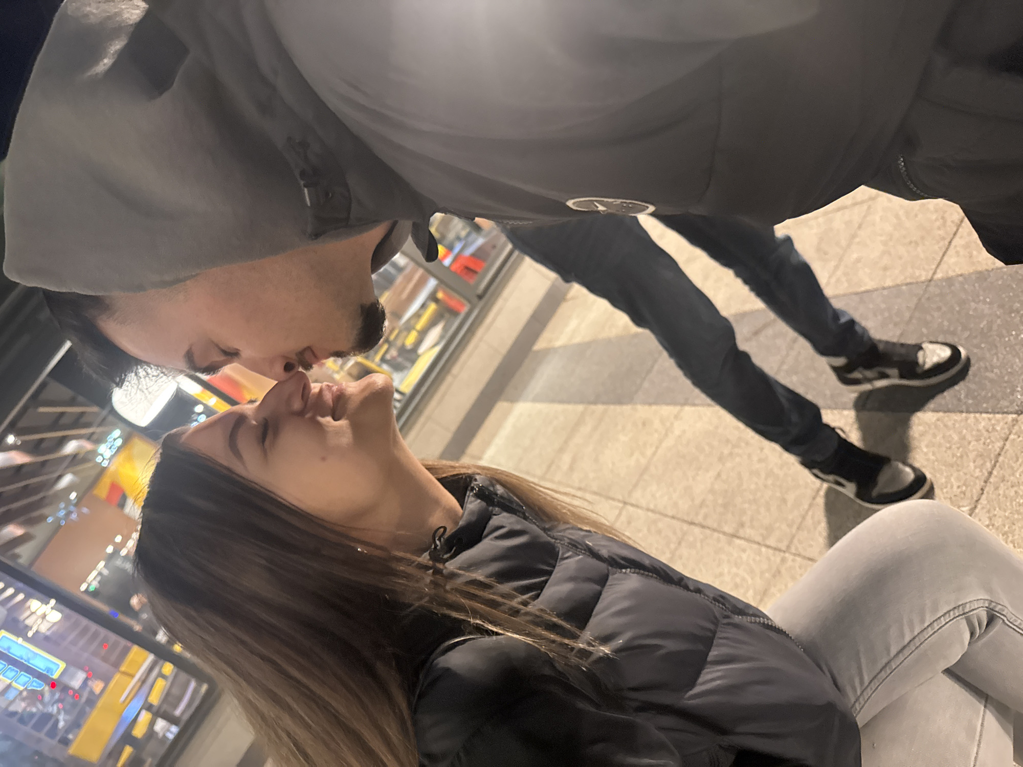 A medium shot shows a couple, possibly kissing, in what appears to be a public location. The woman is positioned in the lower half of the frame, wearing a dark, quilted jacket and light-colored jeans. Her dark hair is long and straight. The man, in the upper half, wears a gray hooded jacket and dark jeans. His footwear consists of black and white sneakers. The background suggests an urban setting with lights and building structures, potentially a cityscape at night. The presence of reflective surfaces further indicates an indoor or sheltered area. Based on the style of architecture, the location is in the city of Sofia, Bulgaria. The photo is angled, giving a tilted perspective.