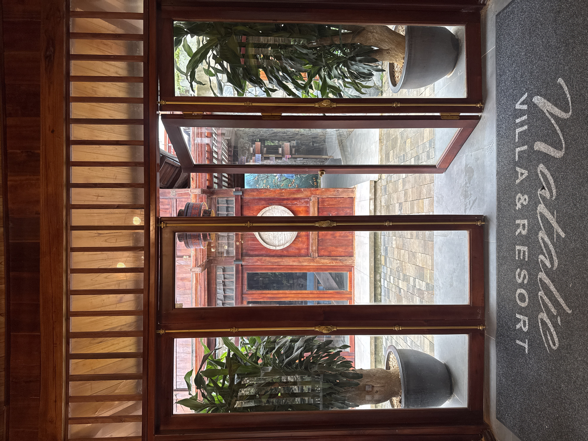 An interior view of a building entrance in Moc Chau, Vietnam, features a double doorway with one door ajar, revealing an outdoor courtyard. The doorway is framed by dark wood and accented with brass hardware. Two large potted plants, one on each side of the entrance, flank the doorway, their lush green leaves contrasting with the warm wood tones. The plants are housed in dark, round planters. Beyond the entrance, the courtyard displays stone paving and traditional Vietnamese architecture, characterized by red wooden structures and ornate details. A welcome mat in the foreground reads "Natalie Villa & Resort" in white lettering. The lighting suggests it is daytime, with natural light filtering through the translucent panels above the doorway.