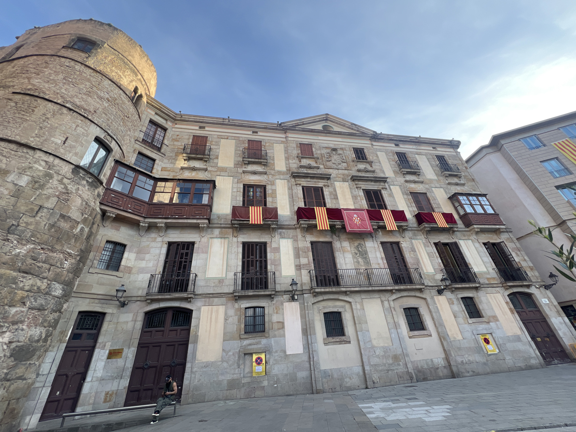 The image depicts a street scene in Barcelona, Spain, featuring historic buildings. On the left stands a stone tower, potentially part of a medieval structure. Adjacent to the tower is a multi-story building with ornate balconies and windows, adorned with Catalan flags, suggesting it may be a government building or cultural center. A person is seated on a bench in front of the building. The environment suggests an urban setting with cobblestone streets. The sky is partially clear.