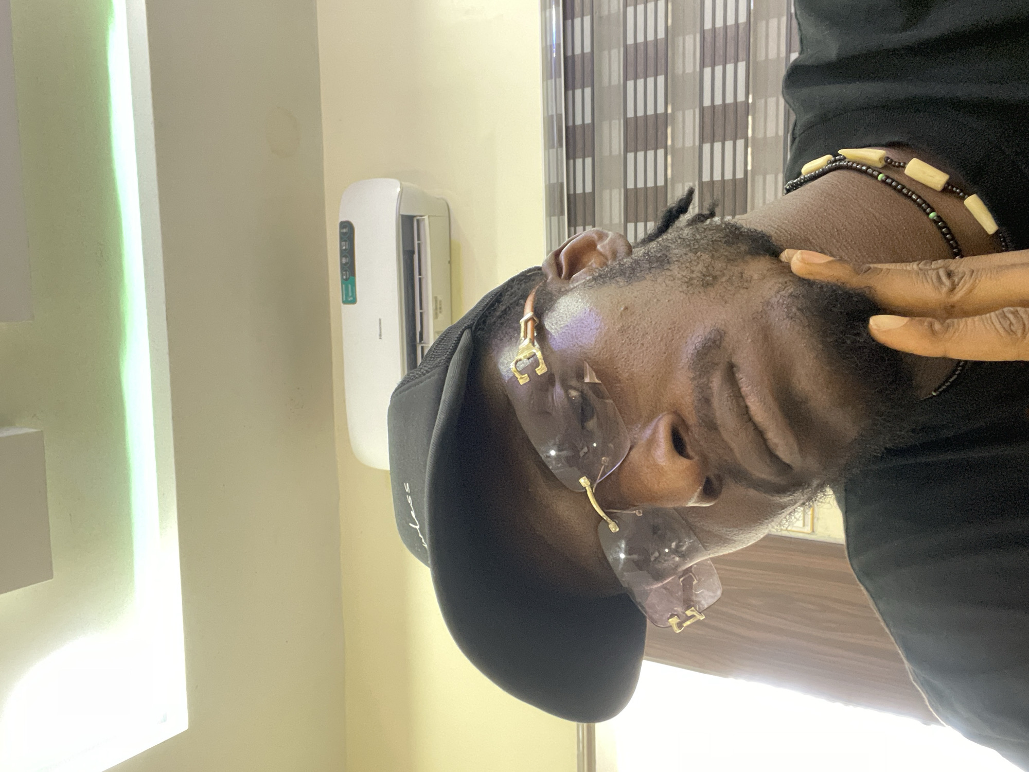 A close-up portrait shows an individual with dark skin, wearing a black baseball cap and clear, rimless sunglasses with prominent gold-colored hardware. The person has a beard or stubble and appears to be smiling or making a relaxed expression, with their head tilted slightly. One hand is visible, gently touching the side of their head. The person is also wearing multiple beaded bracelets on their right wrist, featuring both small black and green beads, and larger off-white/beige tube-shaped beads. A gold-colored earring, possibly shaped like a 'C' or horseshoe, is visible in the left ear.
The setting appears to be indoors, likely a room in Gwarinpa, Nigeria. In the background, to the left, a white Hisense split air conditioner unit is mounted on a light-colored wall. A vertical strip light, possibly an LED fixture, emits a greenish-white glow from the left side of the frame. Towards the upper right, a window with patterned blinds or curtains is visible, suggesting daylight filtering in. The overall mood is casual and relaxed, captured in a close-up, slightly tilted perspective. The only clearly visible text is "Hisense" on the air conditioner unit.