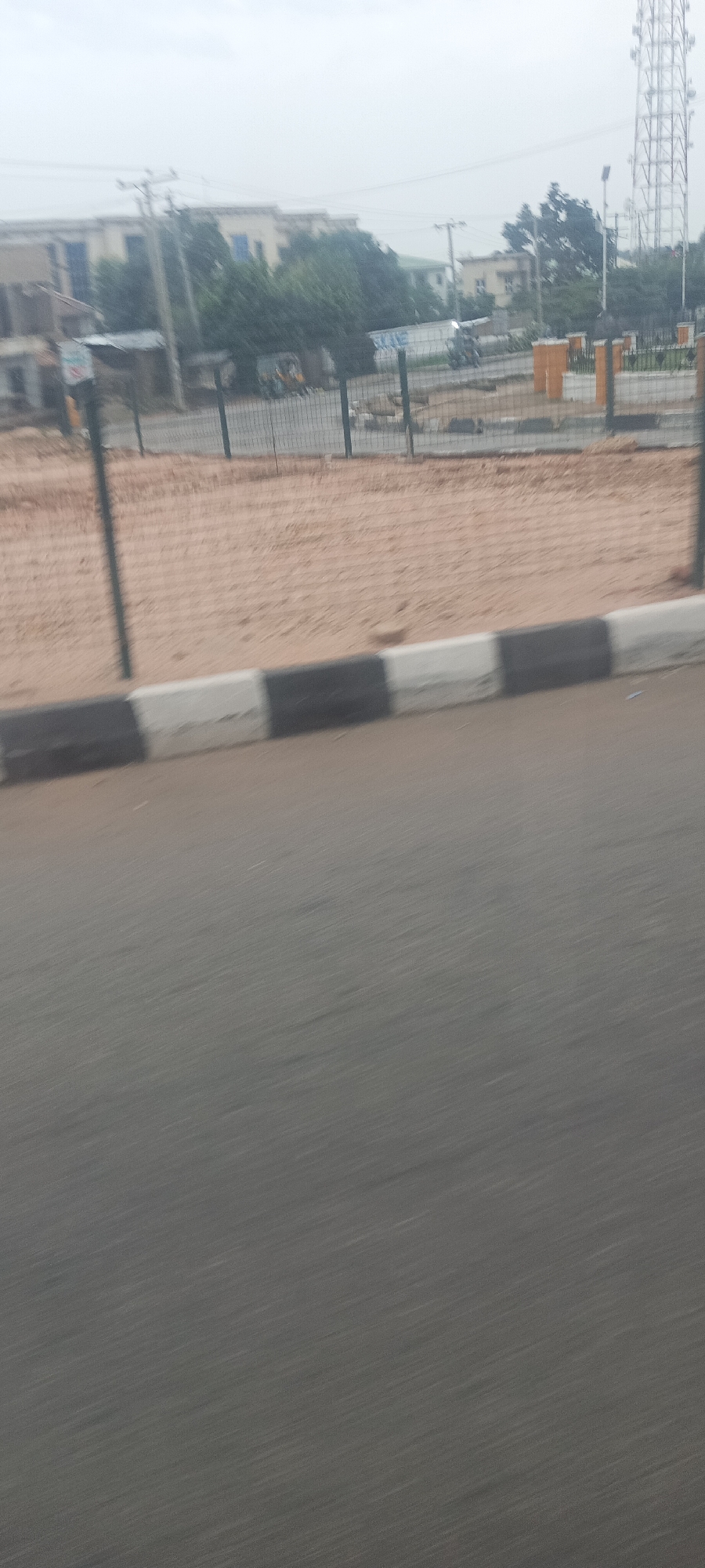 The image captures a street scene in Jos, Nigeria, taken from a moving vehicle under an overcast sky. The foreground features a dark grey asphalt road, heavily motion-blurred, suggesting movement. Above the road, a concrete curb painted with alternating black and white segments separates the roadway from an adjacent area.
Behind the curb, a wide expanse of reddish-brown sandy or dirt ground is visible, stretching towards a dark metal fence that runs across the midground. Beyond this fence, the background reveals a blend of urban infrastructure and natural elements. Several light-colored buildings, some with blue windows or features, rise above a scattering of green trees and bushes. Utility poles with overhead wires are present throughout the background. A prominent metallic lattice communication tower stands tall on the far right.
In the mid-background, a three-wheeled vehicle, likely a Keke Napep, is partially visible on the left side behind the fence. Towards the right, a motorcycle can be seen near what appears to be another street or paved area. This area also features some orange-colored pillar-like structures and a white sign with blue text, where the word "SUKANE" is partially legible. The diffused light and uniform grey sky indicate a cloudy day. No individuals are clearly identifiable in the scene.