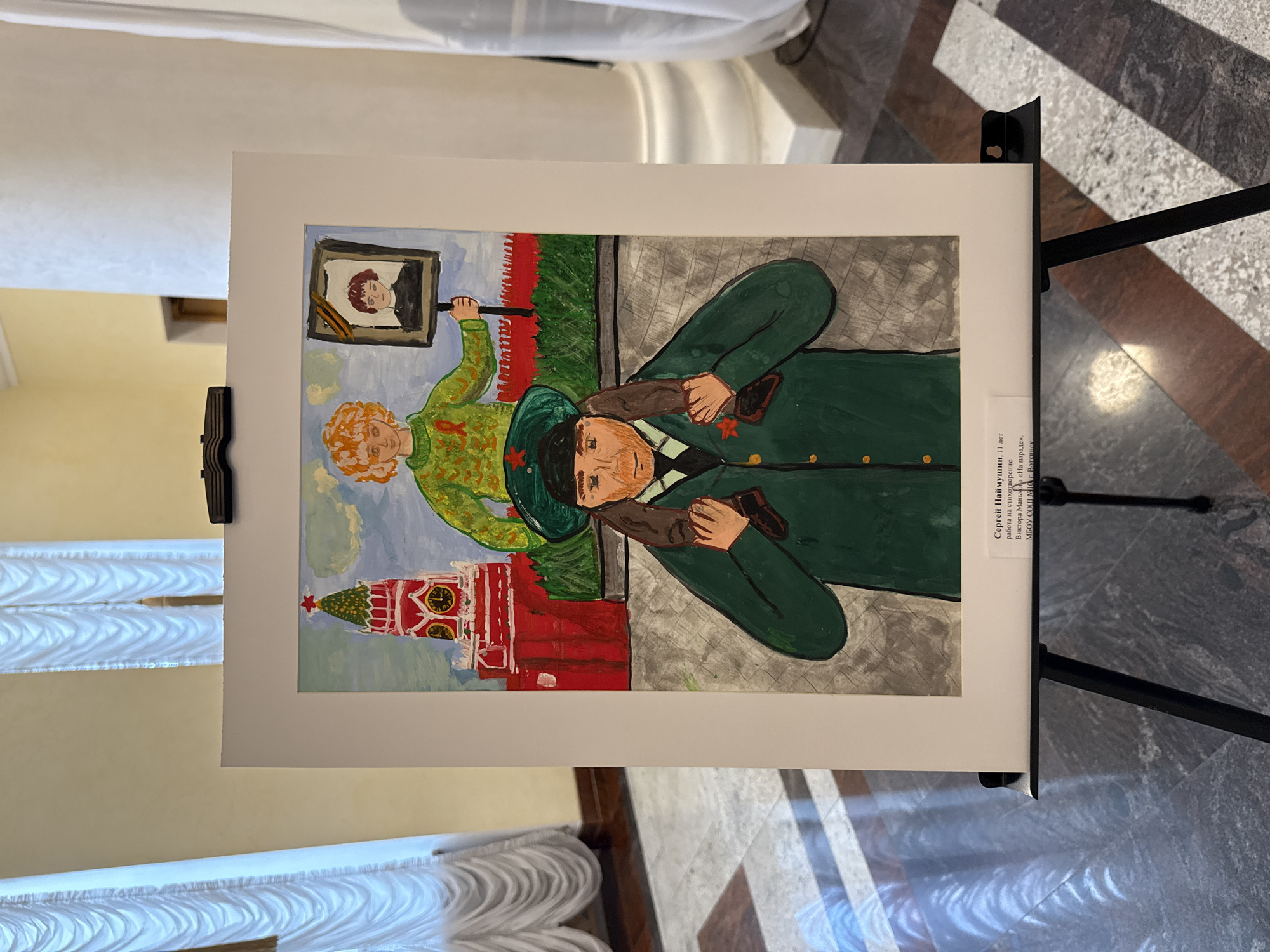 A child's artwork, displayed on a black easel, shows a man in a green military uniform with red stars on his collar and hat, giving a shoulder ride to a child. The child holds up a framed picture of a person. In the background, a depiction of a building resembling the Kremlin is visible. The artwork is identified as "Sergei Naimushin, 11 years old, work on the poem by Viktor Mankov "At the parade." MBOU SOSH No. 10, Votkinsk." The photo is taken indoors, likely in a gallery or exhibition space.