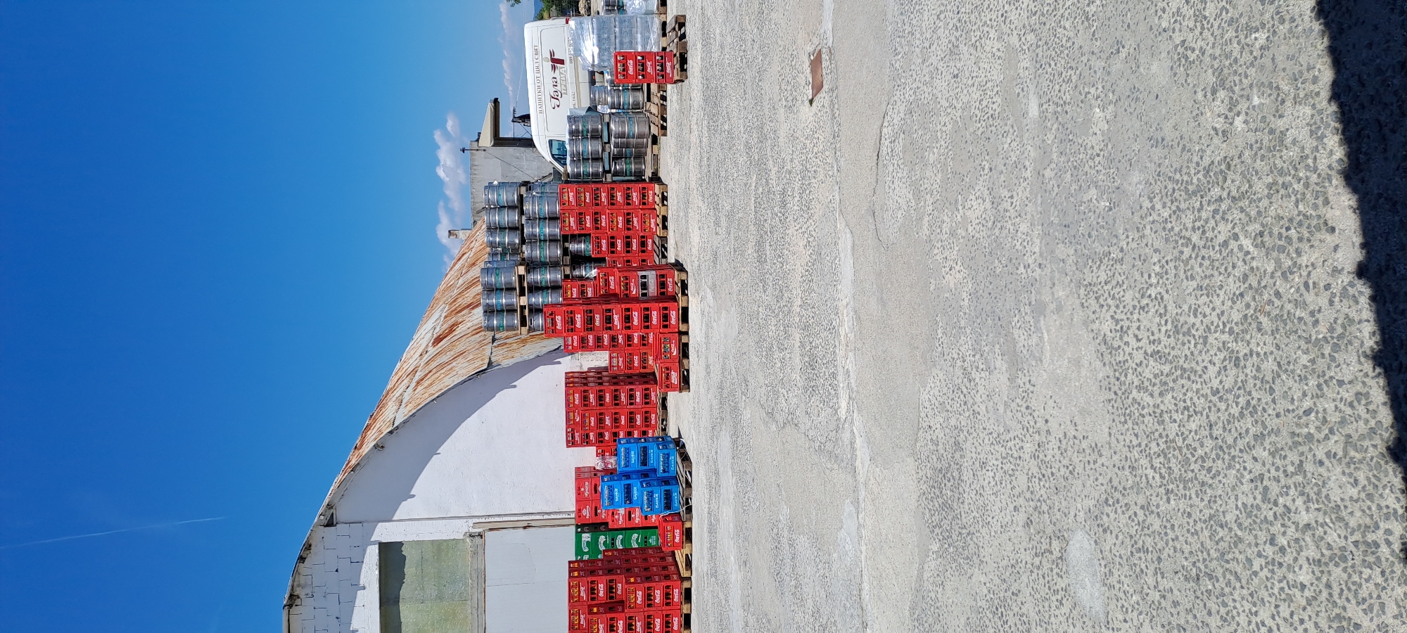 The image shows an outdoor storage area in Velika, Bulgaria, with a white building with a rusty corrugated metal roof on the left. In front of the building, there are stacks of red and blue plastic crates, some of which contain bottles. There are also stacks of metal kegs on pallets. A white van with a logo is parked to the right of the kegs. The ground is a concrete surface with visible cracks and patches. The sky is clear and blue with a single contrail visible. The scene appears to be a storage yard for beverages, likely a brewery or distribution center.