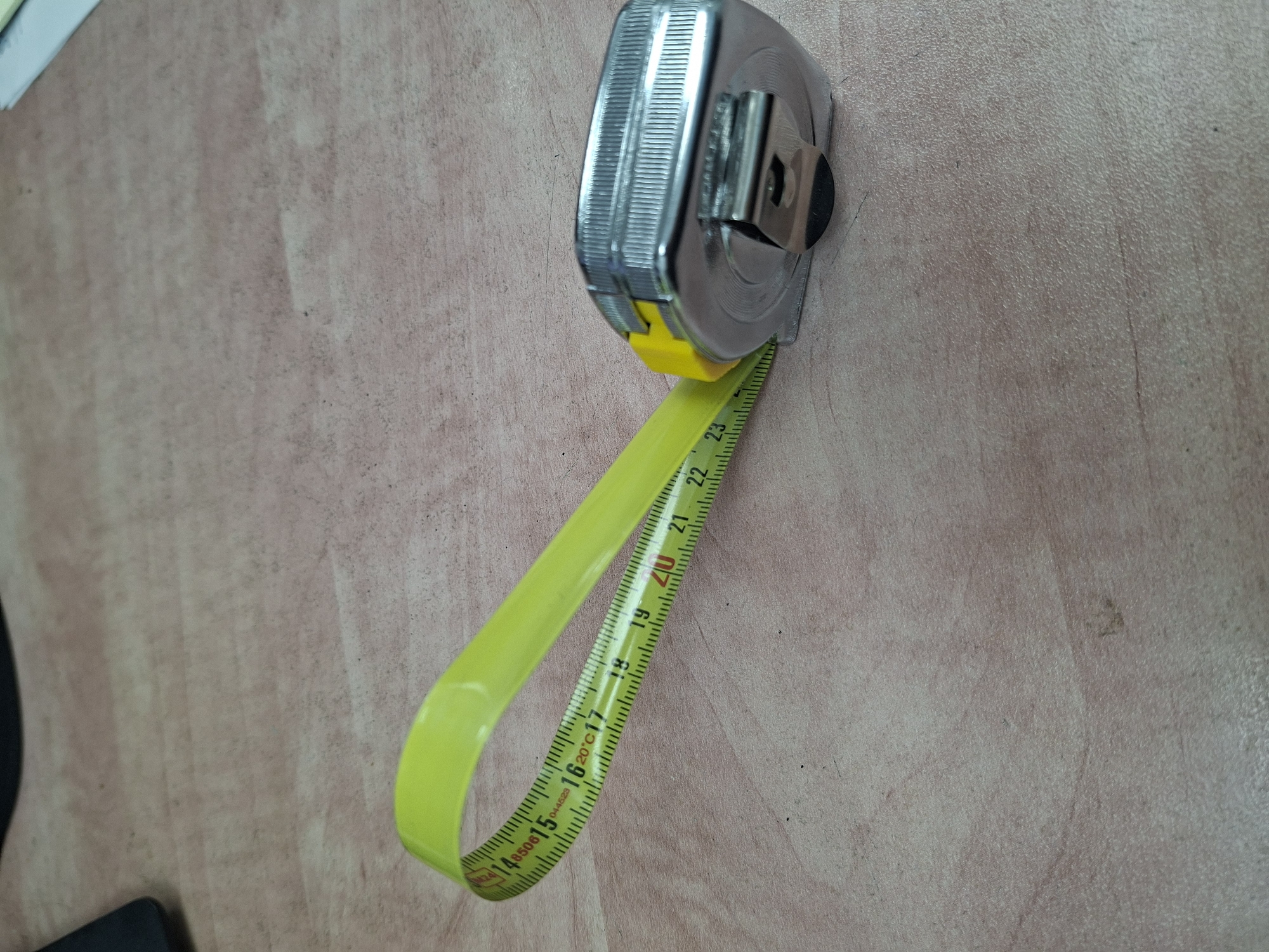A close-up, eye-level shot shows a yellow metal tape measure lying on a light-colored wooden surface. The tape measure is extended and curved, with the metal casing of the tape measure visible on the right side of the frame. The tape itself has black markings and numbers, with the numbers 14, 15, 16, 17, 18, 19, 20, 21, 22, and 23 clearly visible. The wooden surface appears to be a tabletop with a visible grain pattern. The scene is well-lit, with soft shadows. There are no people or other objects clearly visible in the frame. The context of the location in the city of Shomron Regional Council, Palestinian Territories is not discernible from the image itself.