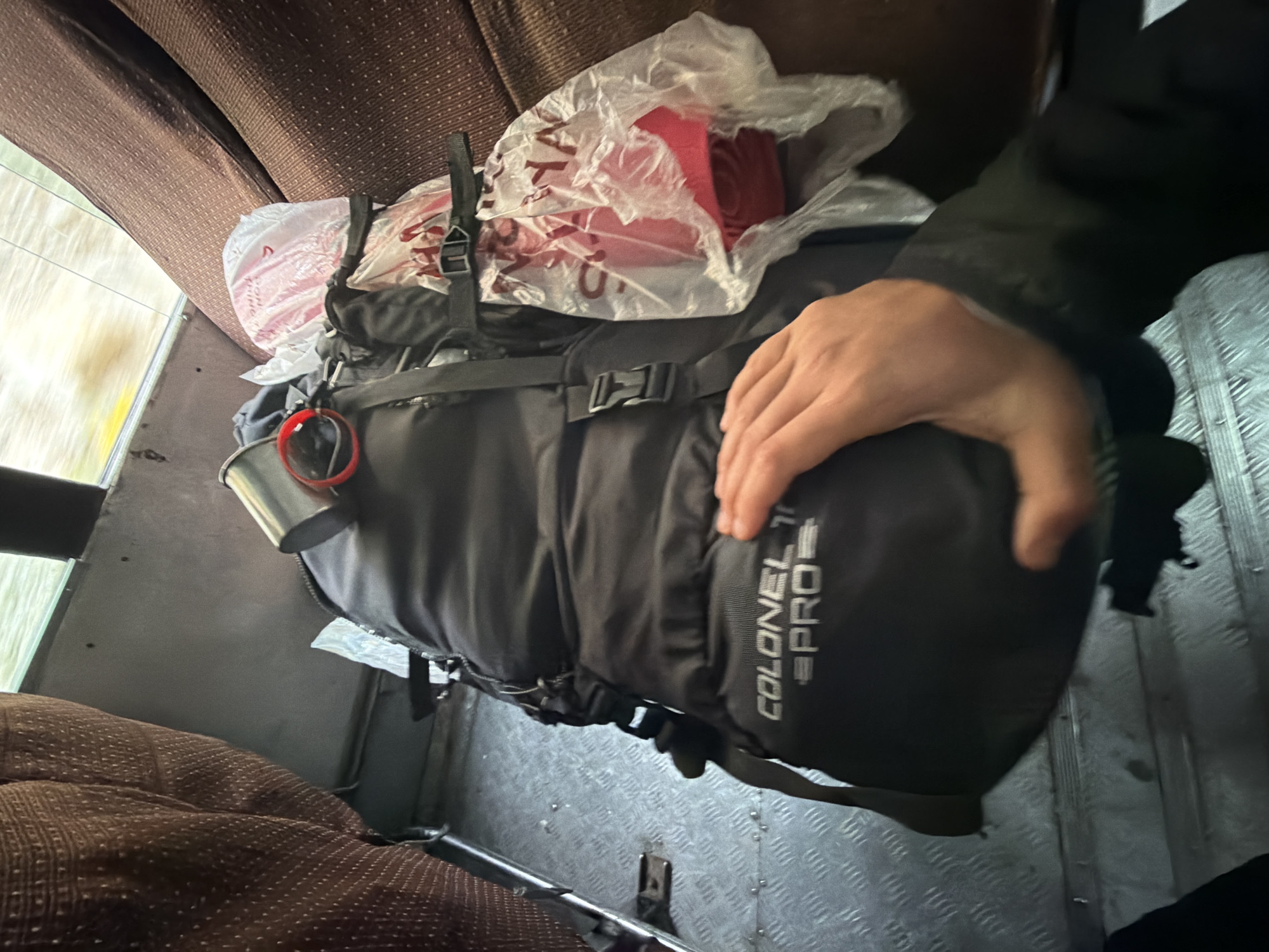 A person's hand rests on a black backpack with "COLONEL PRO" visible on the front. The backpack is situated on the floor of what appears to be a vehicle, possibly a bus, with brown fabric seats visible on either side. A metal cup with a red strap is attached to the top of the backpack. To the right of the backpack, a clear plastic bag contains red items. The floor of the vehicle is made of metal with a diamond plate pattern. The background outside the window is blurred, suggesting movement. There is no discernible information about the city of Hymia, India in the image.