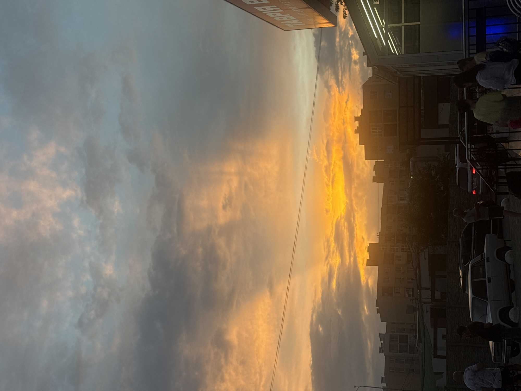 This image captures a sunset over the city of Avanhard, Ukraine. The sky is a dramatic canvas of orange and blue hues, with clouds illuminated by the setting sun. In the foreground, several people are visible, some standing and conversing, while others are near parked cars. A white sedan and a dark SUV are present. Buildings, typical of an urban environment, form the backdrop, and a sign with Cyrillic lettering is partially visible on the right side of the frame. The scene suggests a late afternoon or early evening setting.