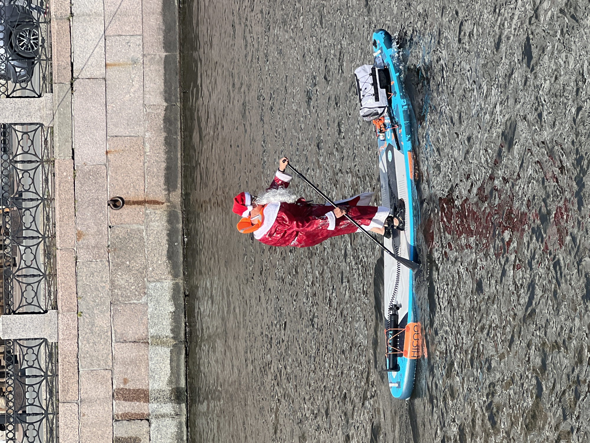 A person dressed as Santa Claus is paddleboarding on a river in Saint Petersburg, Russia. The person is wearing a red suit with white trim, a Santa hat, and a white beard. They are holding a paddle and are standing on a blue and white paddleboard. The paddleboard has a bag and some other equipment on it. The river is wide and the water is dark. In the background, there is a stone embankment with a metal fence and some buildings. The sky is blue and there are some clouds. The scene appears to be festive and unusual, as Santa Claus is not typically seen paddleboarding.