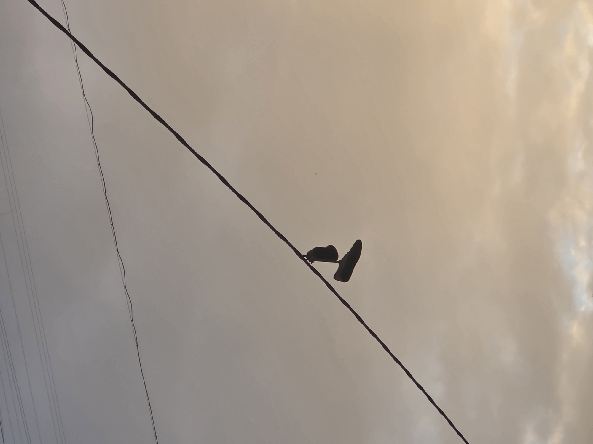 A low-angle shot shows a pair of shoes hanging from a wire. The shoes appear to be dark-colored. The background is a cloudy sky in varying shades of gray, beige, and white. Additional wires are visible in the lower-left corner. The photo was taken in Perm, Russia.