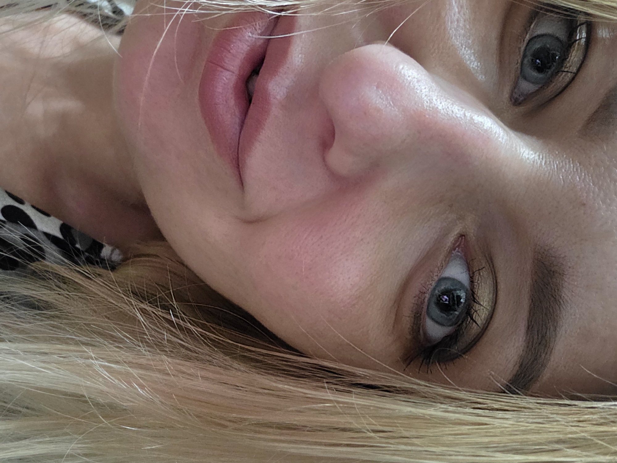 A close-up, high-angle portrait features a female subject with fair skin and light blonde hair. Both eyes are visible and appear light blue or grey. The subject's left eye (viewer's right) is more open, showing dark eyeliner or mascara and golden-toned eyeshadow on the eyelid. The right eye (viewer's left) is partially obscured by the nose and cheek. The nose is prominent in the foreground, displaying visible pores and a slight natural sheen on the skin. The lower lip is visible, slightly parted, with a soft pink or rose hue. Strands of light blonde hair frame both sides of the face. The skin on the face exhibits natural texture with visible pores and a subtle glow, particularly on the cheeks and nose. A black and white patterned garment is visible on the subject's lower right side (viewer's left). The background is entirely out of focus and indistinct, suggesting an indoor setting with soft, ambient lighting. There are no discernible elements within the image that indicate a specific geographic location such as Minsk, Belarus.