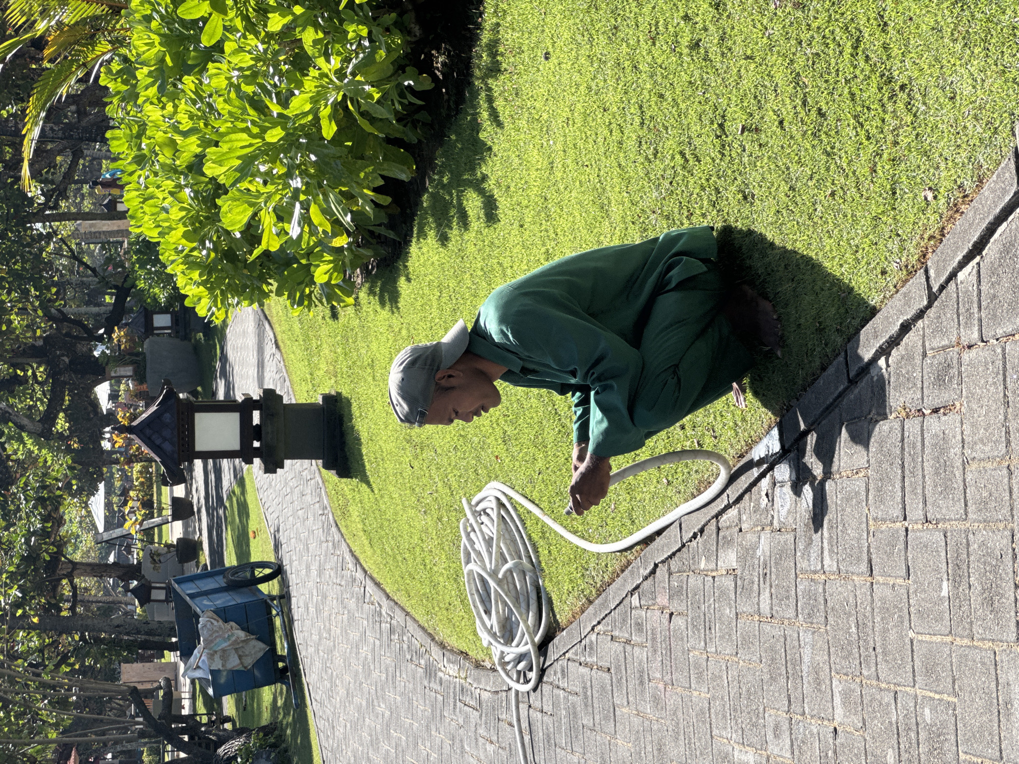 A person in green work clothes and a grey cap is crouching on a paved walkway in Kuta Selatan, Indonesia. They are holding a white garden hose and appear to be attaching or adjusting a nozzle. A coiled length of the same hose lies on the walkway next to them. To the left of the person is a green lawn bordered by a row of trees and a garden light. Further to the left, there is a blue cart partially visible among trees and other garden features. The paving stones of the walkway show signs of sun and shadow, and the overall scene is bathed in sunlight.