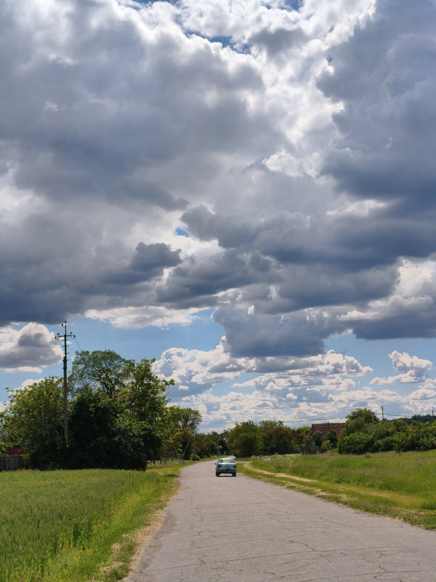 The image depicts a rural road leading towards a light-colored car in Ustyvytsia, Ukraine. The road, cracked and showing signs of wear, is flanked by grassy verges and vegetation. To the left, a telephone pole stands beside green trees and fields. The right side features similar greenery, with glimpses of building roofs suggesting the presence of a settlement. Above, the sky is filled with a mix of dark storm clouds and patches of blue sky with white clouds.