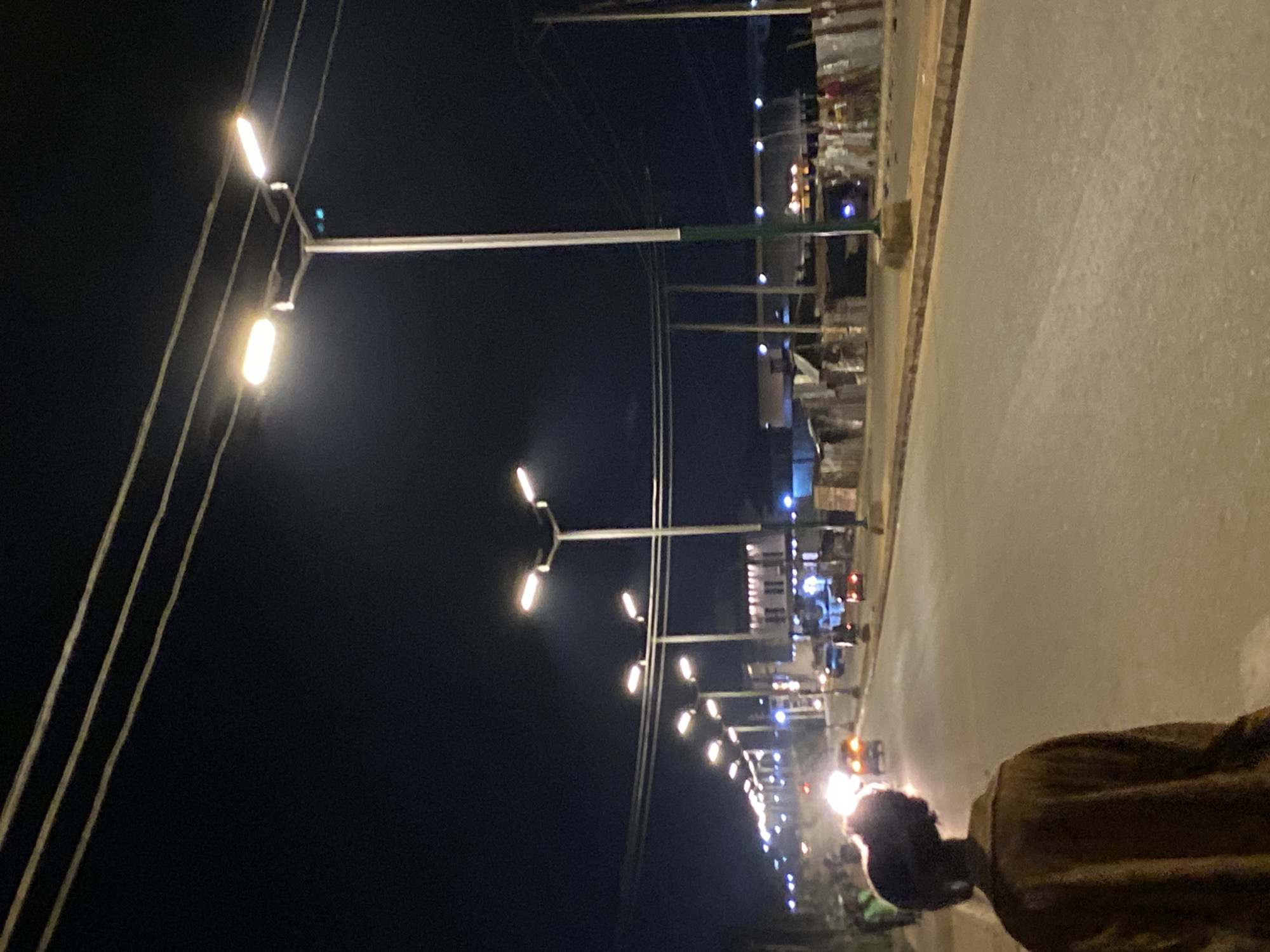 The image depicts a nighttime scene along a street in Bukuru, Nigeria. A series of bright, modern streetlights illuminate the dark environment, extending into the distance along the roadside. Some streetlights have multiple lamps, casting a significant glow. Overhead, power lines or cables are visible against the dark sky.
In the bottom right foreground, a person is seen from behind, wearing a dark or brown jacket. Their head is silhouetted against the bright headlight of a vehicle, likely a motorbike, positioned further down the road. The paved road surface occupies the right side of the frame, reflecting some of the artificial light. Faint outlines of buildings or market stalls can be observed on the left side of the street in the background, also marked by various small lights. The overall atmosphere suggests a typical town street at night with ongoing activity, though details are softened by the darkness. No text is visible in the image.