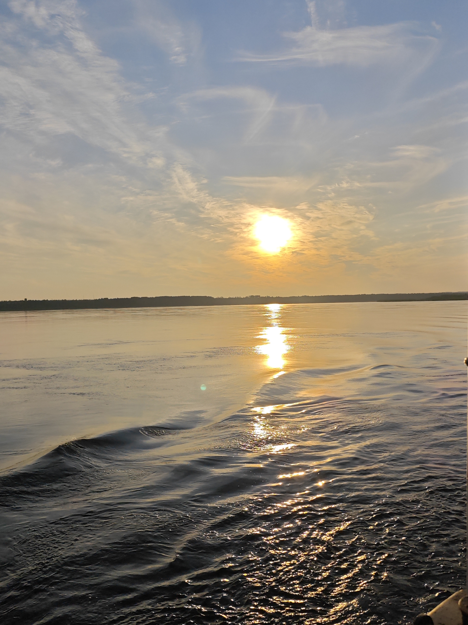 A vertical, eye-level, full shot captures a sunset over a wide body of water. The sun, a bright orb, is positioned in the upper center of the frame, casting a golden reflection on the rippling surface of the water. Wispy clouds streak across the pale blue sky, tinged with hues of orange and yellow near the horizon. A dark, silhouetted tree line marks the distant shore, suggesting a rural or undeveloped area. The foreground is dominated by the dark, choppy water, with small waves and a wake trailing from the perspective of the viewer, indicating movement. No people or structures are visible in the scene.