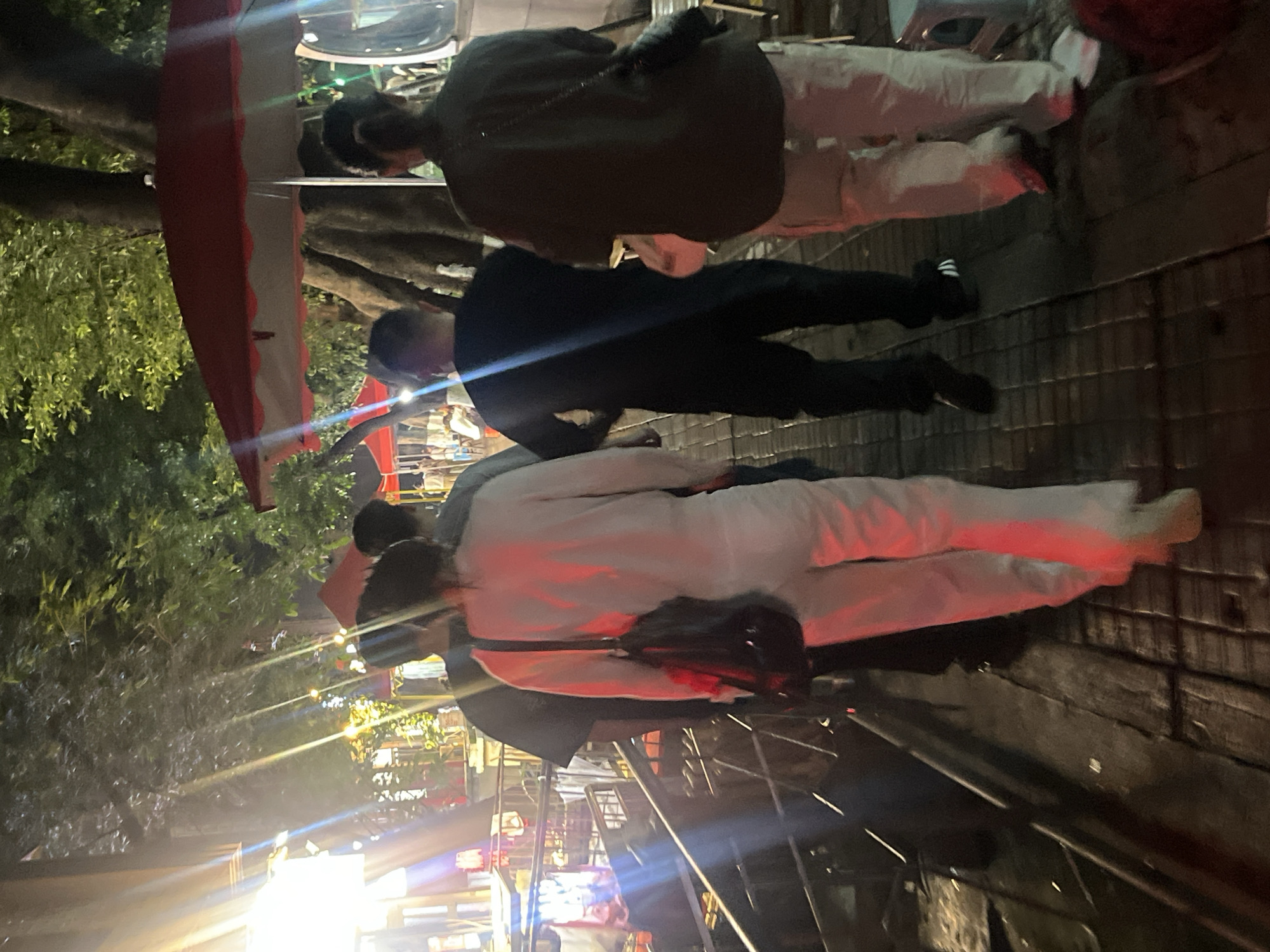 Four individuals are walking on a wooden boardwalk at night. The boardwalk is illuminated by artificial lights, some of which cast strong reflections. Trees with green foliage are visible in the background. Red and white umbrellas are also present. The individuals are dressed in casual clothing. One person in the foreground is wearing a light-colored outfit and carrying a black bag over their shoulder. Another person is wearing a black t-shirt and dark pants. A third individual is wearing a dark shirt and light-colored pants, with a black bag slung across their chest. The fourth person is partially obscured. The environment suggests an outdoor market or street scene.