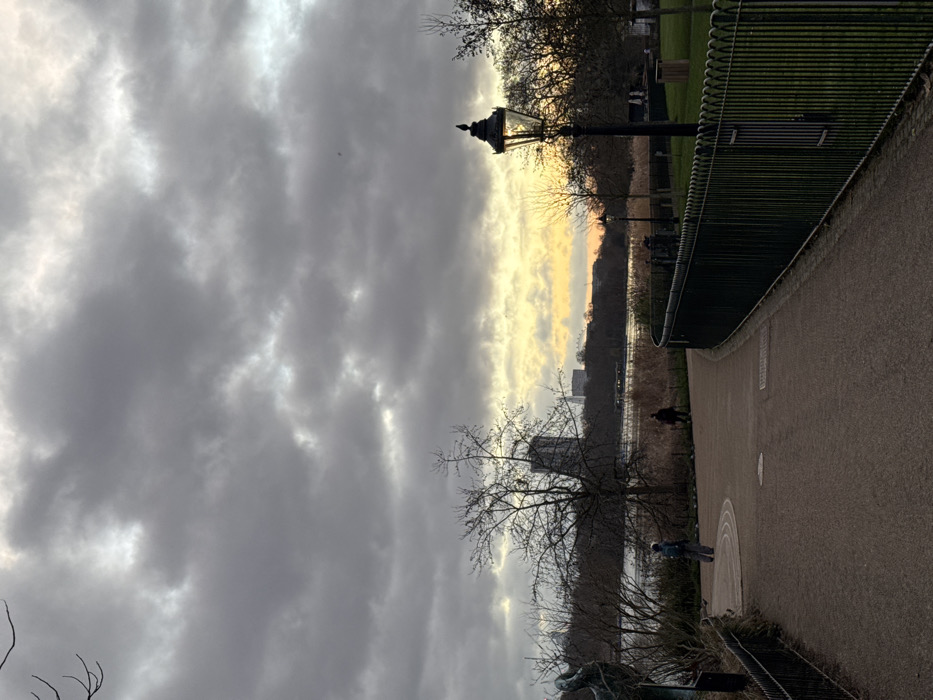 This image depicts a late afternoon or early evening scene within Hyde Park, London, England, looking east-southeast across the Serpentine lake towards the central London skyline. The sky is largely covered by heavy, dark grey clouds, with a distinct band of yellow-orange light visible on the horizon, indicative of sunset.
In the foreground, a wide, dark-colored paved pathway curves gently to the right. On the immediate right, a dark green ornamental metal fence with a curved top separates the path from a grassy area. A black, illuminated Victorian-style street lamp stands prominently just behind this fence on the right, with a small dark bird perched on its finial.
Two individuals are visible on the path. One, dressed in a blue jacket and light pants, stands facing away from the viewer on the path's left side. Further down the path, a second individual in dark clothing walks towards the right, appearing smaller due to distance. To the far left, a dark, abstract sculpture or tree base is partially visible.
The midground features the calm, dark surface of the Serpentine lake, reflecting the subdued sky. The lake's edge is bordered by tall, dry reeds and numerous bare-branched deciduous trees, suggesting late autumn or winter.
In the background, on the far bank of the lake, a dense line of bare trees forms the distant horizon. Above these trees, the central London skyline is discernible, including a tall, dark building and the distinctive, illuminated structure of the London Eye Ferris wheel, silhouetted against the bright yellow-orange sky.