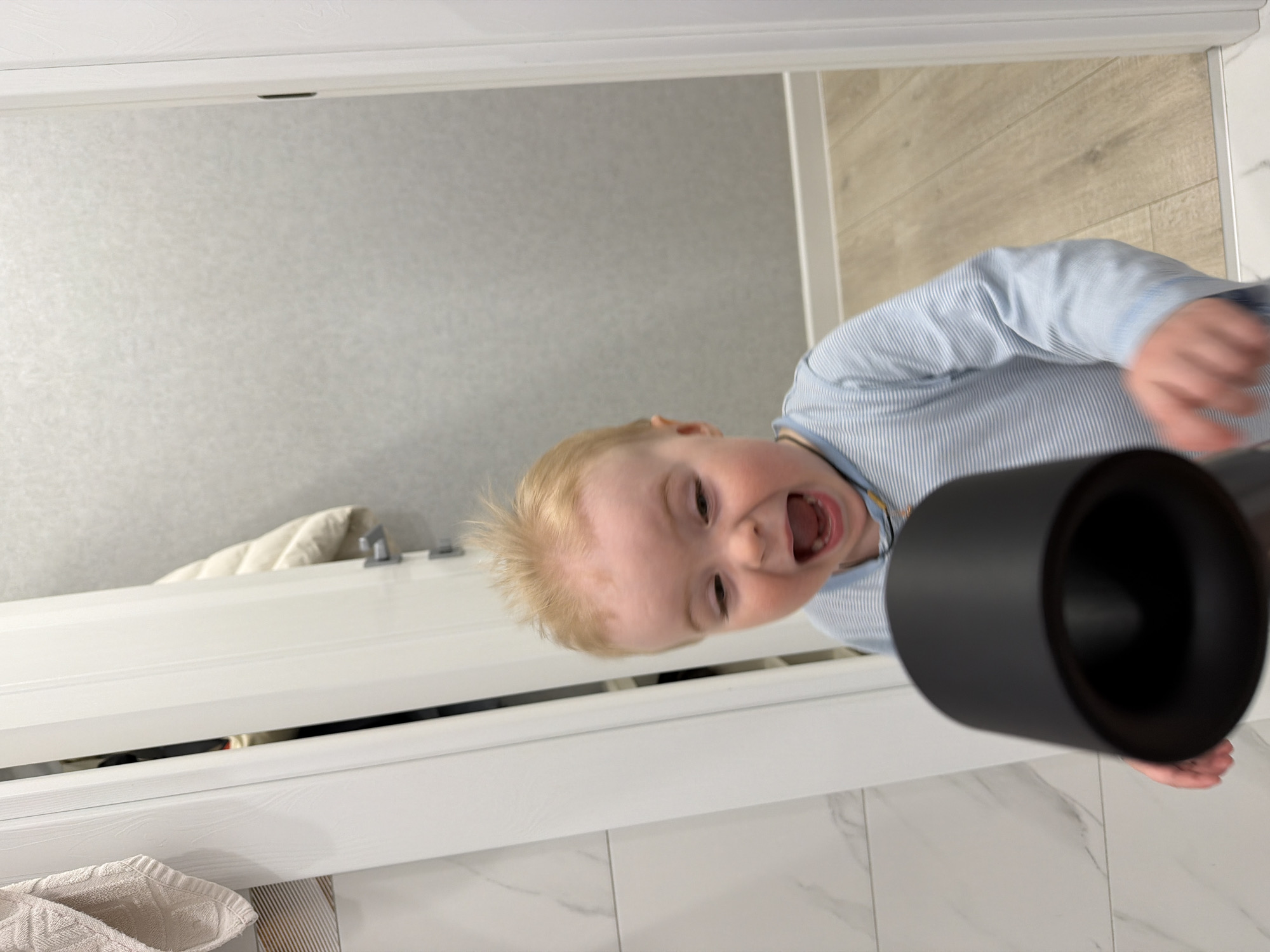 A low-angle, indoor shot captures a toddler in Votkinsk, Russia, as they peer through a doorway. The child has fair skin, blond hair, and wears a light blue and white striped long-sleeved shirt. The toddler's mouth is open in a yell or laugh. A black cylindrical object is held in front of the camera, slightly obscuring the child. The background includes a door frame, a gray wall, and a tiled floor. The doorway leads into a space with a white bathtub and a folded white towel.