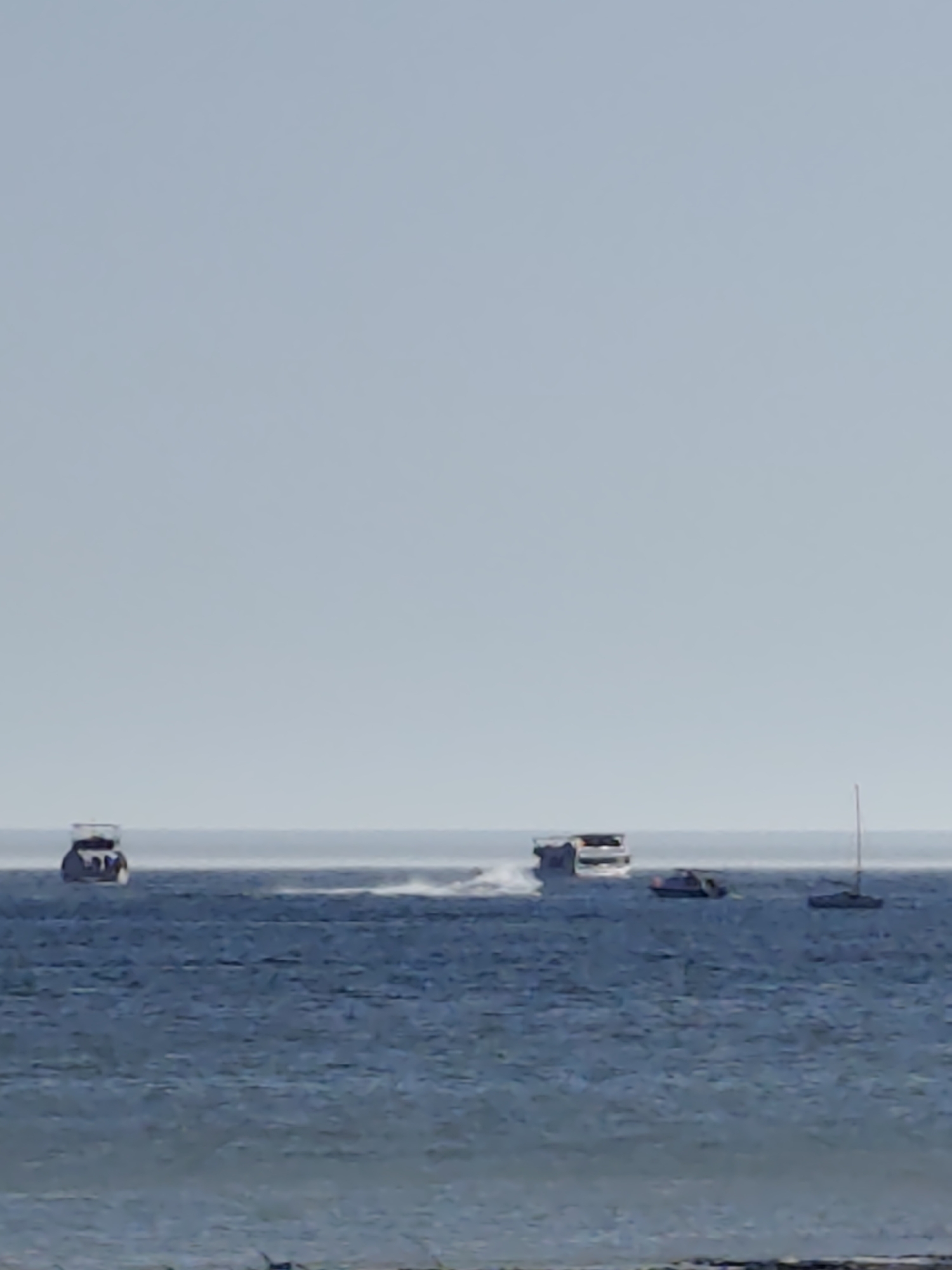 This is a photograph of the Caspian Sea, likely taken from a beach area, with boats on the water. The water is choppy and a deep blue. The sky is a pale, clear blue, with a thin line of lighter blue or white visible on the horizon, suggesting a distant shoreline or haze.
In the foreground, there are blurry hints of what could be a rocky or sandy beach. The main focus is on the water and the vessels.
From left to right, the following can be observed on the water:
* A small white boat with a dark top, appearing to be a pleasure craft, is positioned on the left side of the frame.
* Further to the right and slightly further from the camera, there is a larger, multi-decked boat that is creating a significant wake, suggesting it is moving at speed. It appears to be a tour boat or ferry.
* To the right of the larger boat, there is a smaller dark-colored vessel, possibly a RIB or a small speedboat.
* On the far right, a sailboat with its mast clearly visible is anchored or drifting.
The scene is tranquil and appears to be under daylight conditions with clear weather. The location is the Caspian Sea, and given the description of Aktau, Kazakhstan as the requested location, this photograph is likely depicting a scene along the coastline of Aktau.