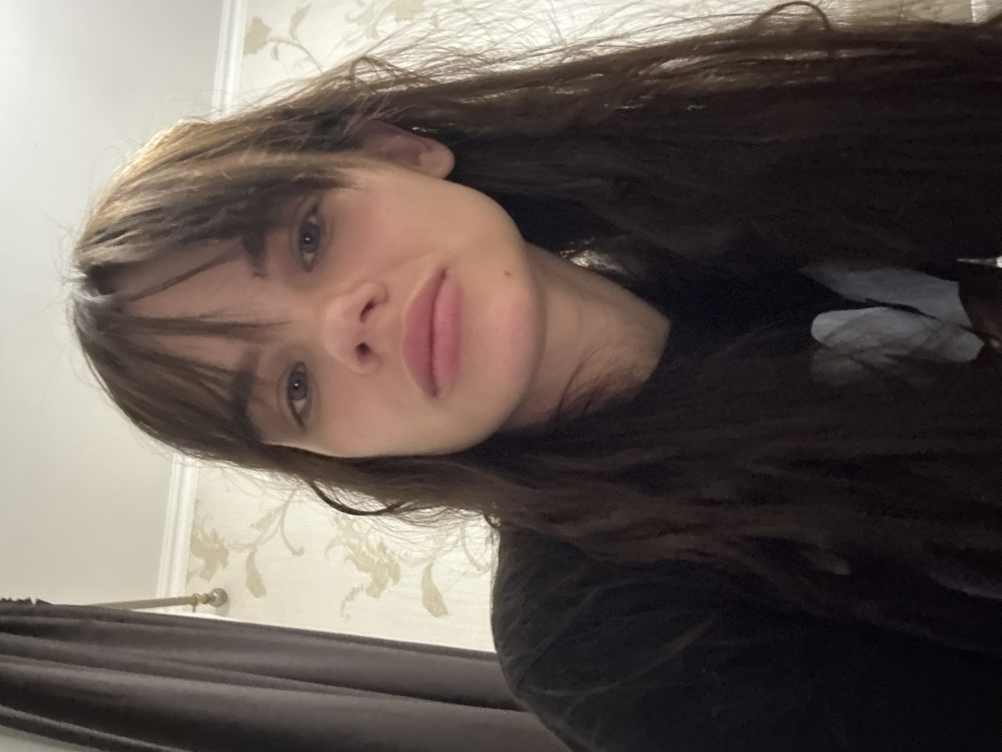 A young individual with long, dark brown, wavy hair and bangs is captured in a close-up, angled indoor portrait. The person, likely female, has light-colored eyes and full lips, displaying a relaxed or slightly pouting expression directly at the camera. They are wearing a dark-colored top, possibly black. The setting appears to be indoors in Rostov-on-Don, Russia. The background features a light-colored wall with subtle floral or leaf-patterned wallpaper and a white decorative molding. Dark brown curtains and a gold-colored curtain rod finial are visible on the left side of the frame. The image is a selfie, taken from a low and tilted angle, focusing on the subject's face and upper body. The lighting is soft, evenly illuminating the face. No visible text is present.