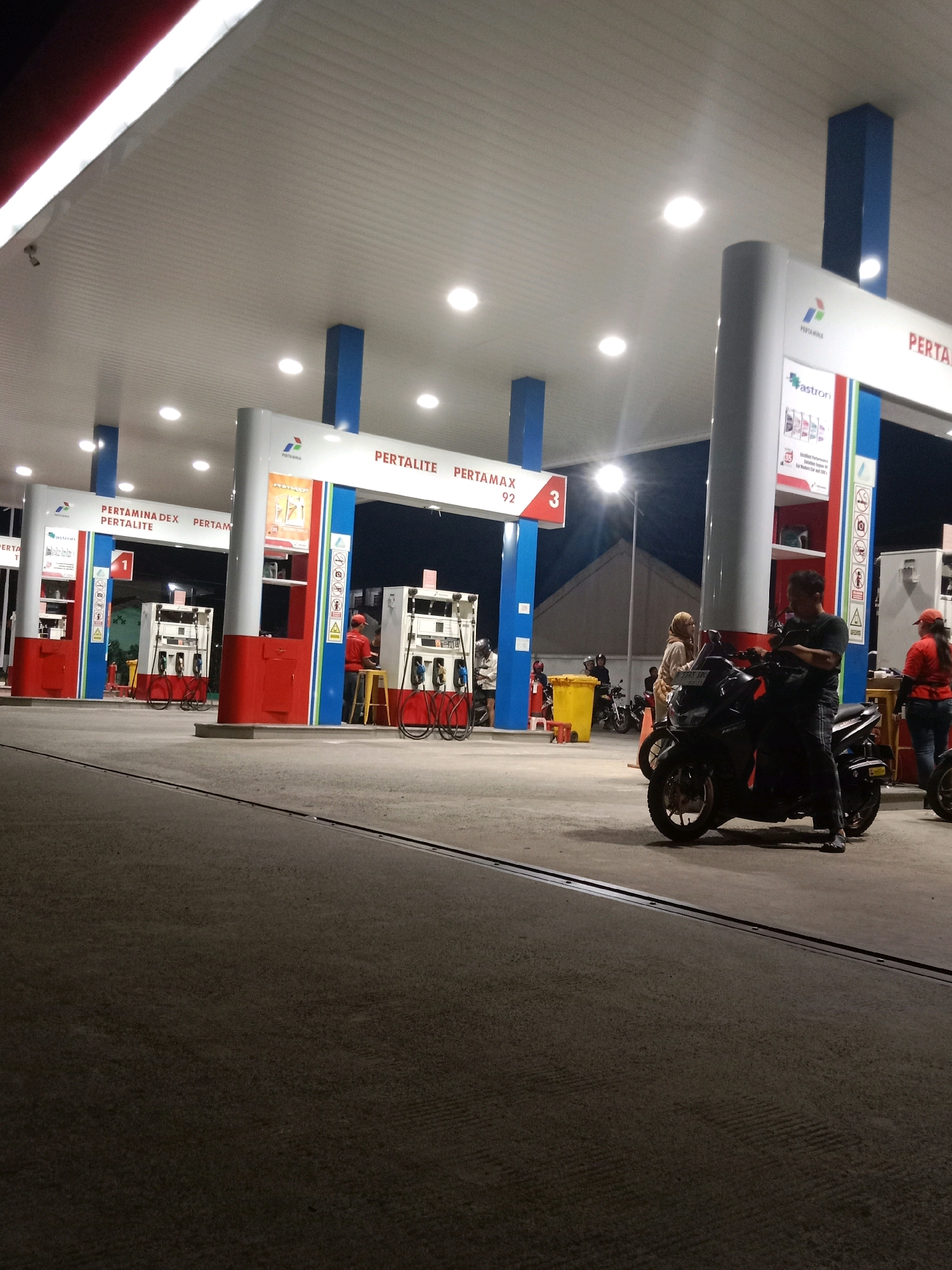 This image captures a night scene at a Pertamina gas station in South Tangerang, Indonesia, featuring multiple fuel dispensers under a covered canopy. The canopy is illuminated by overhead lights, casting a bright glow on the concrete ground. Several Pertamina-branded signs are visible, advertising different fuel types such as "PERTAMINA DEX PERTALITE" and "PERTAMAX 92." Two fuel dispensers are prominent in the foreground, with hoses and nozzles readily accessible. A man in a red uniform, likely a gas station attendant, is standing by one of the dispensers, appearing to interact with a customer. A man on a black motorcycle is parked at a dispenser, and another person is seated on the motorcycle. In the background, other motorcycles and a bright yellow trash can are visible. Several individuals are present at the station, some appearing to be customers and others possibly staff. The overall environment suggests a typical night operation at a public fueling station.