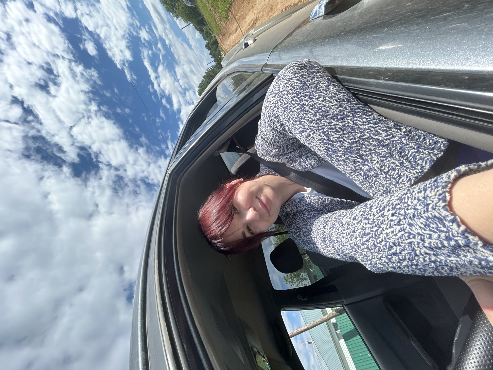 A woman with reddish-brown hair is taking a selfie from the passenger seat of a car. She is leaning out the open window with her arms resting on the car door. She is wearing a blue and white knitted sweater over a white t-shirt. The car appears to be on a dirt road in a rural area. In the background, there are trees, a green fence, and a house with a white roof. The sky is blue with white, fluffy clouds. The scene suggests a road trip or travel outside of a city environment.