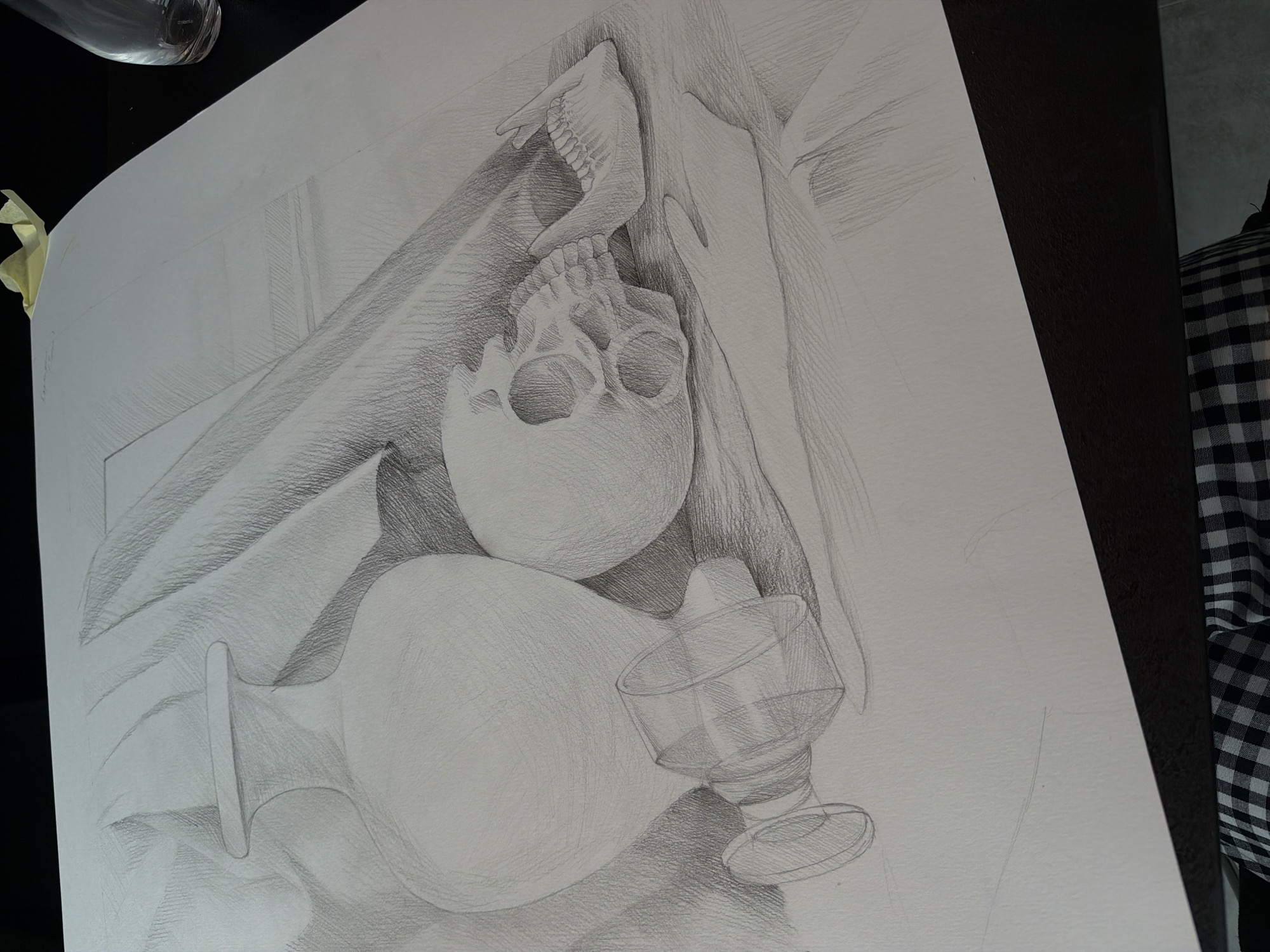The image depicts a detailed pencil drawing of a still life arrangement. The central subject is a human skull, positioned beneath a jawbone. Below the skull, there is a long glass bottle laid horizontally, with a stemmed glass resting near its base. The drawing is on a large sheet of paper, slightly angled, with a strip of yellow tape visible on the left edge. The background is suggested with shading and implied forms. A dark surface, possibly a desk or table, is visible on the right side of the image, partly covered with a checkered fabric.