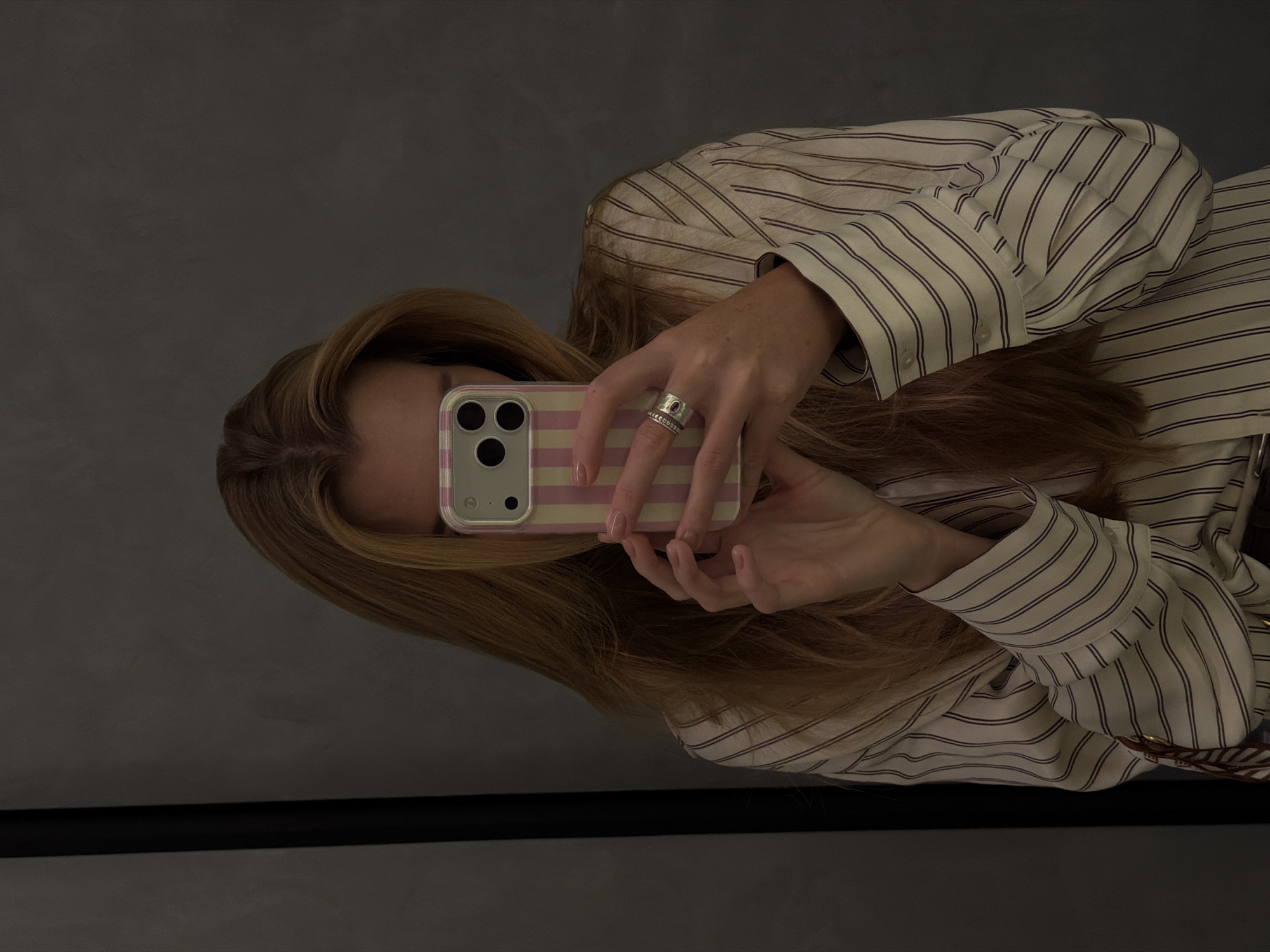 A woman is taking a selfie with a pink and white striped phone case in front of a gray wall. She is wearing a white striped shirt and multiple rings. The location is unknown and cannot be determined to be Bishkek City, Kyrgyzstan from the information available in the image.