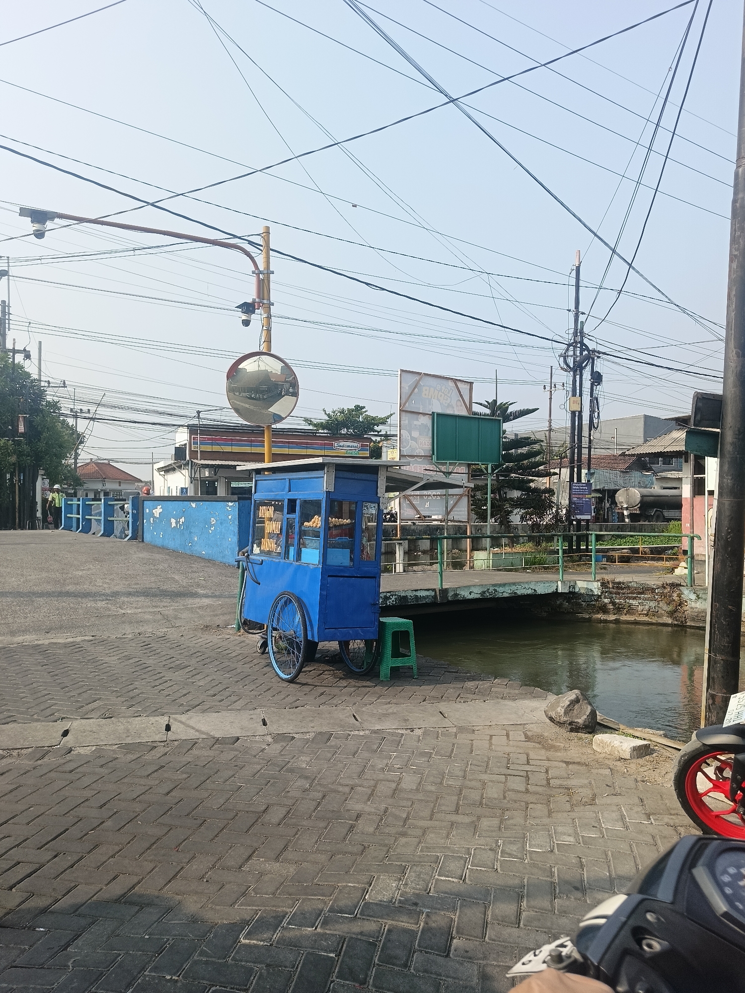 The image depicts a street scene in Wedi, Indonesia, with a bright blue food cart prominently featured in the foreground. The cart, which appears to be selling "Batagor" and "Siomay", has visible food items inside. A green stool is placed next to the cart. The cart is situated on a paved sidewalk made of interlocking bricks, which leads towards a body of water with a small bridge crossing it. The bridge has a green railing and stone supports.
In the background, several buildings are visible, including what appears to be a convenience store with colorful signage, some residential houses with tiled roofs, and a larger building with a white tank truck parked in front of it. Utility poles with a dense network of electrical wires crisscross the sky, indicating an urban or semi-urban environment. A large convex mirror on a pole is positioned near the food cart, reflecting the surroundings. Two individuals wearing light-colored uniforms and dark helmets are standing near a blue wall on the left side of the image. The sky is clear and bright, suggesting it is daytime. A motorcycle is partially visible in the lower right corner of the frame. The overall impression is a typical street scene in a town in Indonesia, with the food cart being a central element of daily commerce and street life.