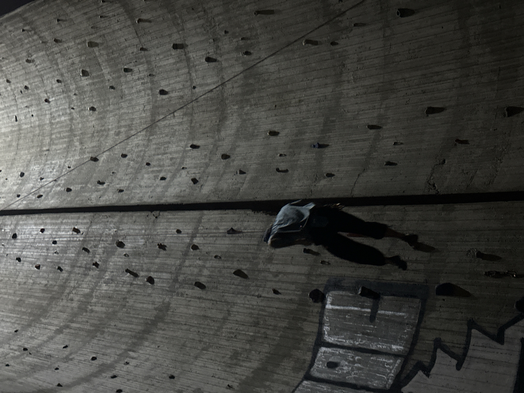 A climber is traversing a concrete climbing wall. The wall is dark gray and has numerous climbing holds attached to it. Graffiti is visible on the lower right side of the wall. The lighting is dim, suggesting it is nighttime. The location is not specified, only "Unknown, Unknown".