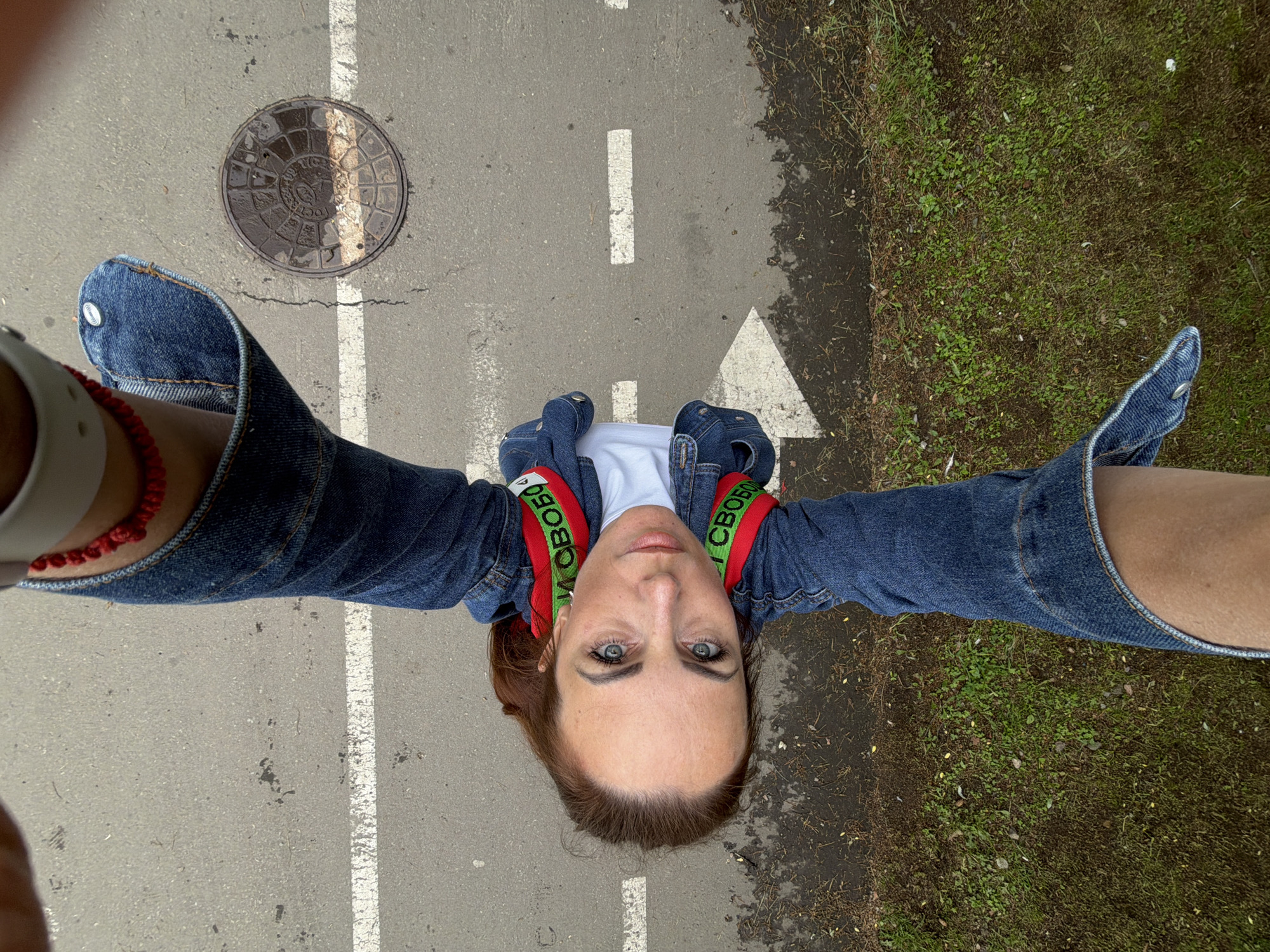 In a high-angle shot, a woman with auburn hair and blue eyes is positioned head-down in the center of the frame. She is wearing a white t-shirt and a denim jacket with red and green striped bands on the sleeves that read "И СВОБОДЕ". A red beaded bracelet is visible on her wrist. Her legs, clad in denim jeans, extend upwards out of the frame, framing her face. She is outdoors, on asphalt pavement marked with white lines, one of which appears to be a directional arrow. A manhole cover is visible to her right. The surrounding area includes a patch of grass and some scattered leaves. The scene does not contain any discernible landmarks or elements specifically identifying it as being in Birobidzhan, Russia.