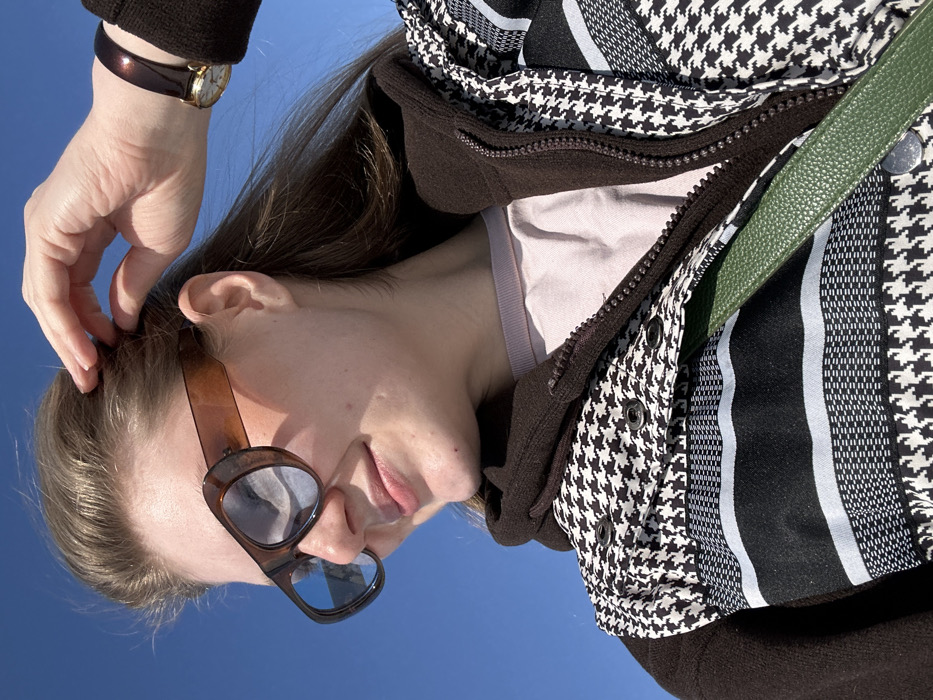 This is a close-up, low-angle portrait of a young woman standing outdoors under a clear blue sky in Новое Шигалеево, Russia. The subject, a woman with light brown hair, is positioned with her head slightly tilted and her gaze directed towards the viewer's right. She is smiling faintly. Her right hand is raised, with fingers gently touching the hair near her right temple. She wears oversized, brown-framed sunglasses with reflective blue lenses. Her attire consists of a light pink or lilac-colored crew-neck shirt, layered under a dark brown, possibly fleece, zip-up hoodie or jacket. Over these, she wears a vest or jacket featuring a prominent black and white houndstooth pattern, interspersed with horizontal black and light grey striped sections. Several dark buttons or snaps are visible on the upper left front of this patterned garment. A dark green, textured bag strap runs diagonally across her chest. A gold-toned watch with a dark brown strap is visible on her right wrist. The background is an unobstructed bright blue sky, indicating clear weather.
