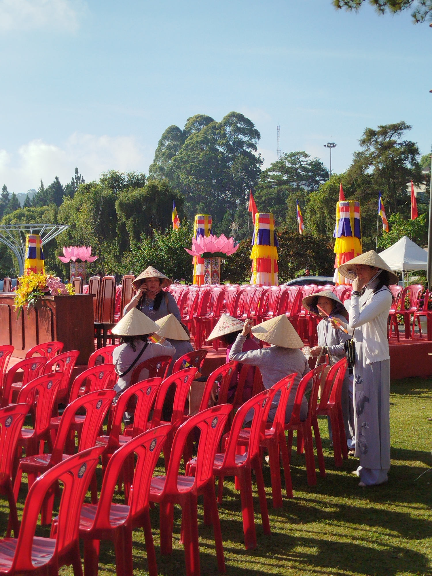 In an outdoor space in Da Lat, Vietnam, rows of red plastic chairs are arranged on a grassy area. Several people wearing traditional conical hats are present, with one standing and holding what appears to be a cigarette. Behind the chairs, there's a raised platform with more chairs and decorations, including colorful columnar structures wrapped in yellow, blue, and red fabric. Pink lotus flower decorations are also visible. Trees and flags are scattered throughout the background, and a clear blue sky is overhead.