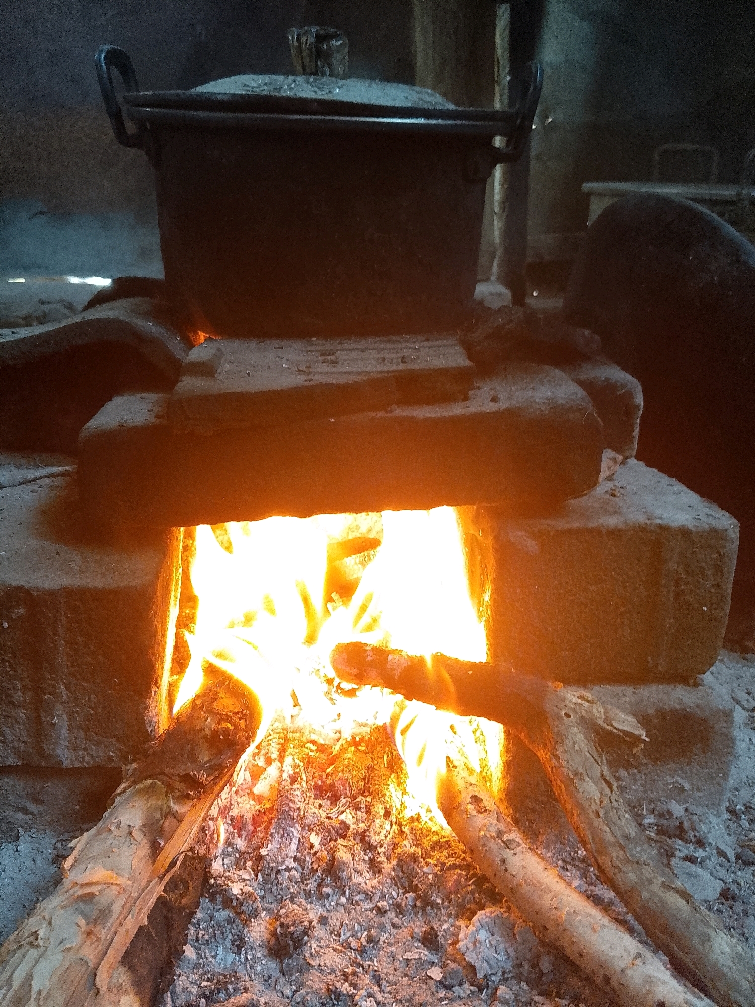 A close-up shot captures a traditional clay stove burning with visible flames and embers, located in Wadang, Indonesia. A dark pot with handles is placed on top of the stove. Several pieces of firewood are arranged within the fire pit, and ash covers the base. The background is dark, suggesting an indoor or dimly lit outdoor setting. The lighting is warm and orange, primarily originating from the fire, highlighting the textures of the wood, ash, and clay. The overall impression is one of simple, rustic cooking.