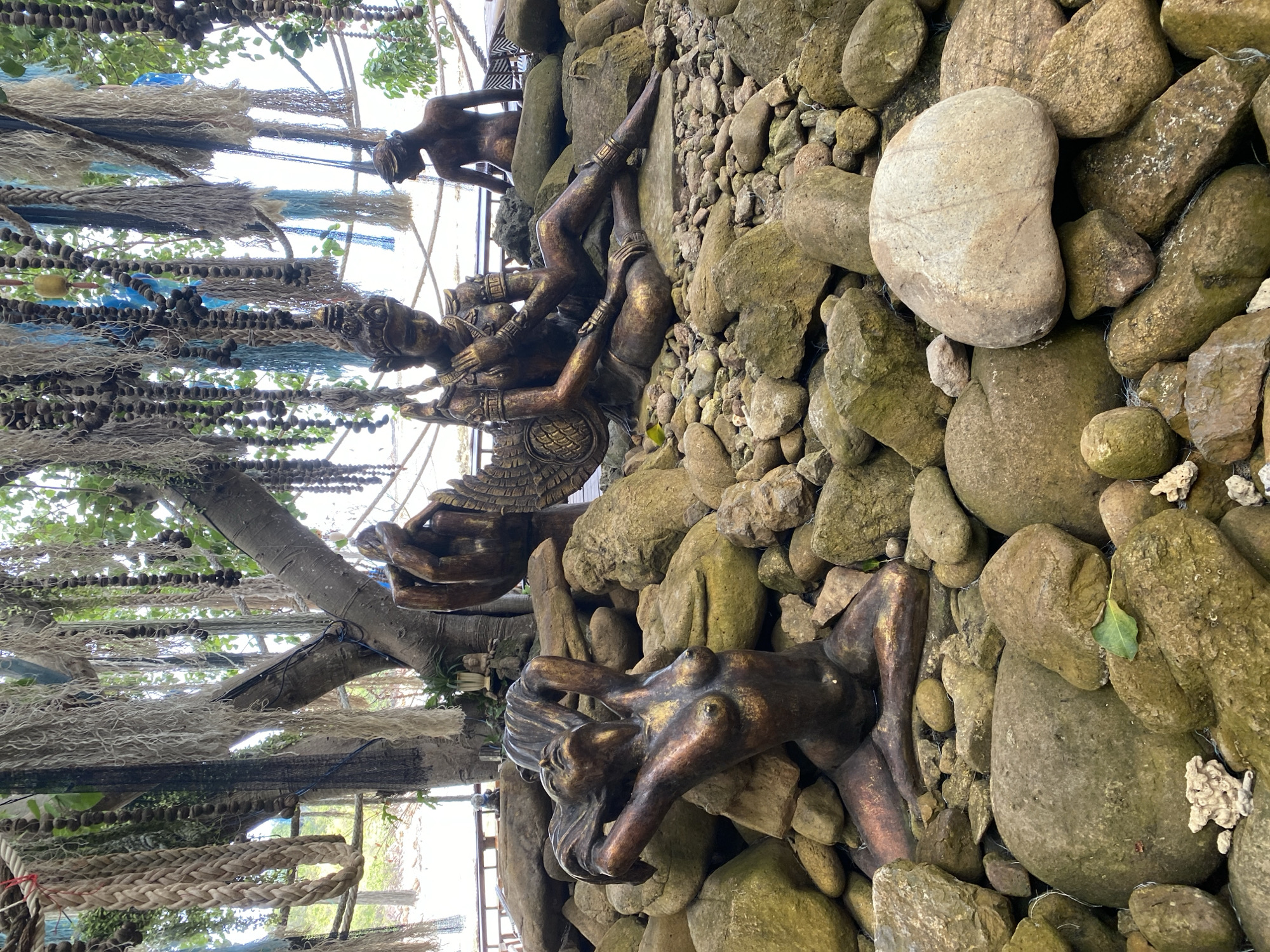 This image depicts an outdoor scene in Koh Phangan, Thailand, featuring several bronze statues situated amongst a collection of large rocks. In the foreground, a female statue is positioned in a reclining pose, with her arms raised and hair cascading down her back. Behind her, another statue of a figure with wings and ornate headdress is seated. In the background, a third statue of a person is visible, facing away from the camera. These statues are surrounded by a variety of rough-hewn rocks, some of which appear to be partially submerged in water. Above the statues, the branches of a tree are adorned with numerous strands of rope and netting, creating a decorative canopy. The overall environment suggests a natural setting with artistic embellishments, possibly a garden or a decorative outdoor space.