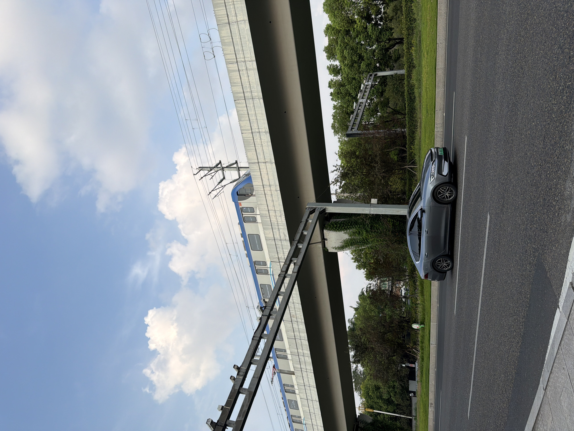A silver sedan is driving on a road in Jiangjin, China. The sedan is in the foreground, and the road is asphalt with white lane markings. In the background, an elevated train track runs above the road. A train is visible on the track, with blue and white cars. Trees and greenery are also visible along the side of the road and beneath the train track. The sky is blue with scattered white clouds.