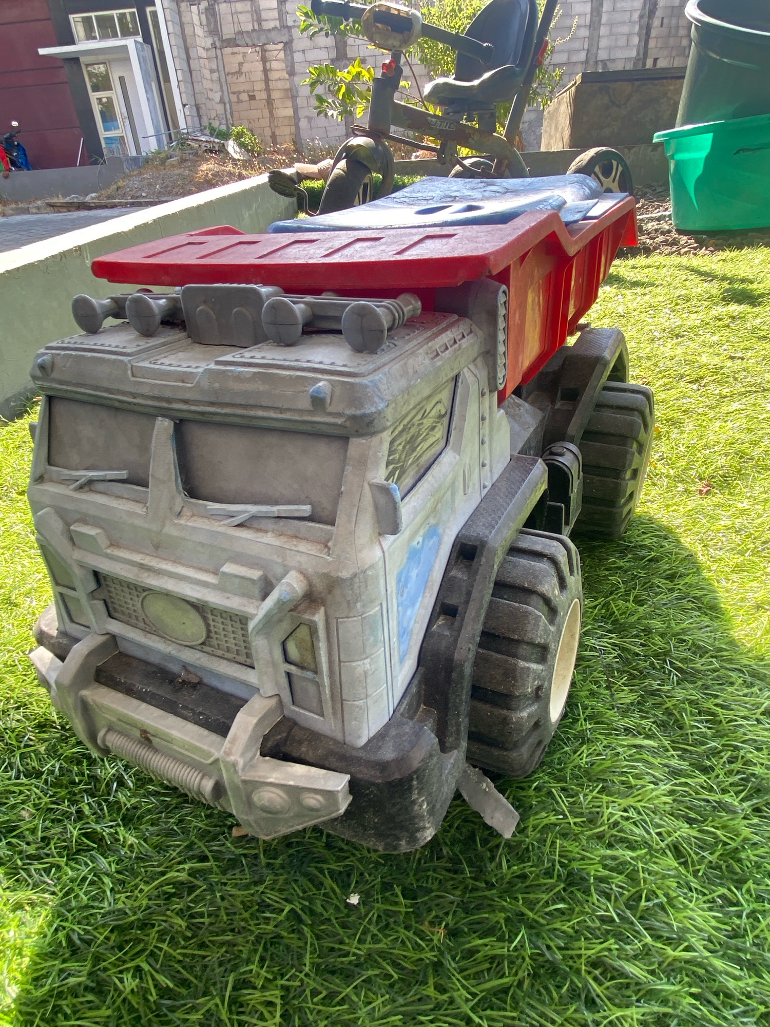 A large, gray, plastic toy dump truck with a red dump bed sits on a patch of green artificial grass outdoors. The truck has four large, black, textured tires and a detailed front grille. In the background, a red motorcycle with a rider in a blue shirt is visible near a building. Further in the background, a brick wall, some trees, and green and black plastic bins are present. The scene appears to be in a residential area.