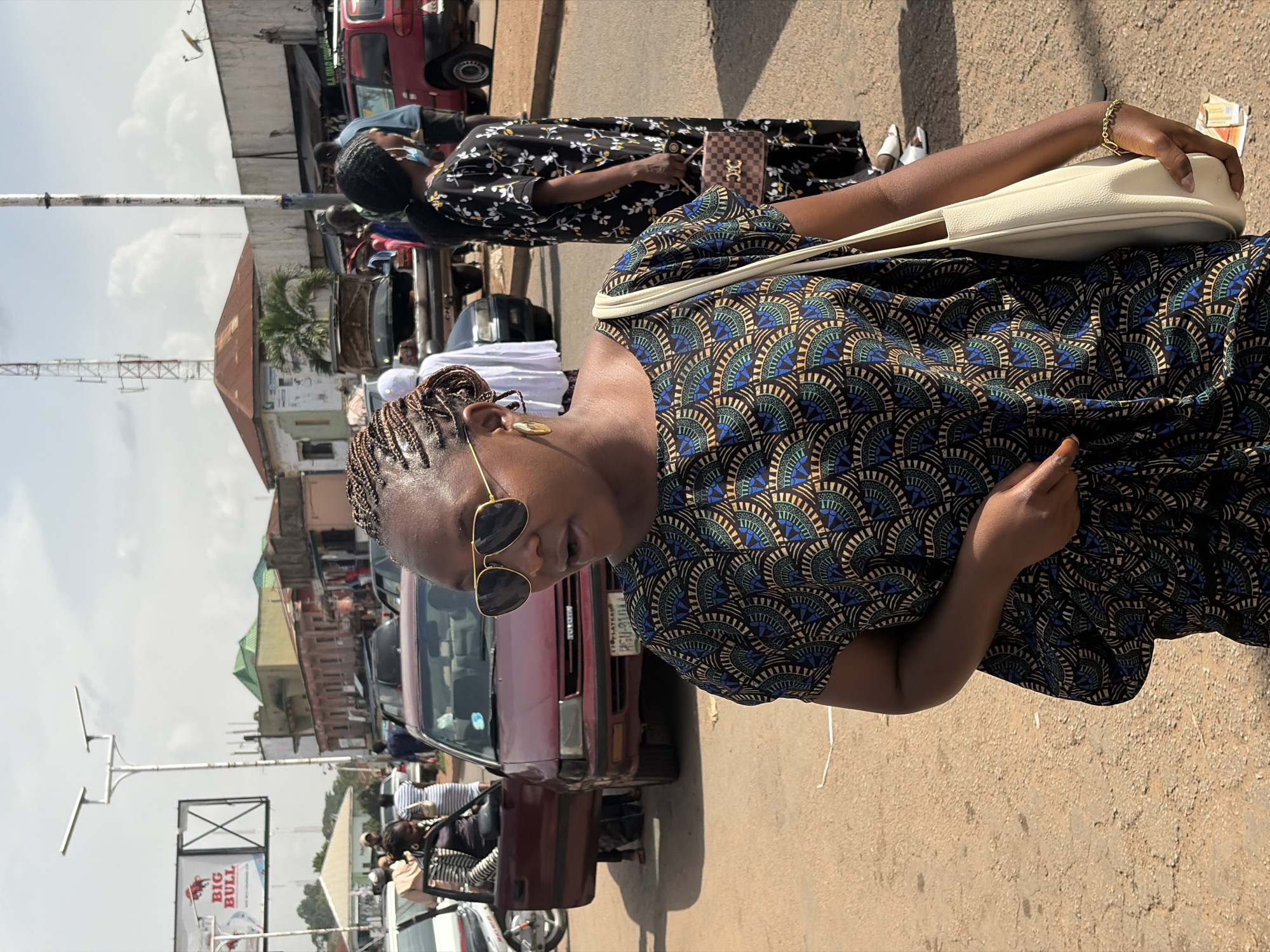 A medium shot captures a woman with braided hair and sunglasses in the foreground, looking to her left with an open mouth. She wears a patterned dress and carries a cream-colored bag over her shoulder. Behind her, a dark red car with an open driver's side door is parked on a dusty road. Another woman in a long black and white floral dress stands to the right, holding a checkered purse. In the background, buildings, a billboard reading "BIG BULL," streetlights, and vehicles suggest an urban environment, possibly in Jos, Nigeria. The sky is partly cloudy.