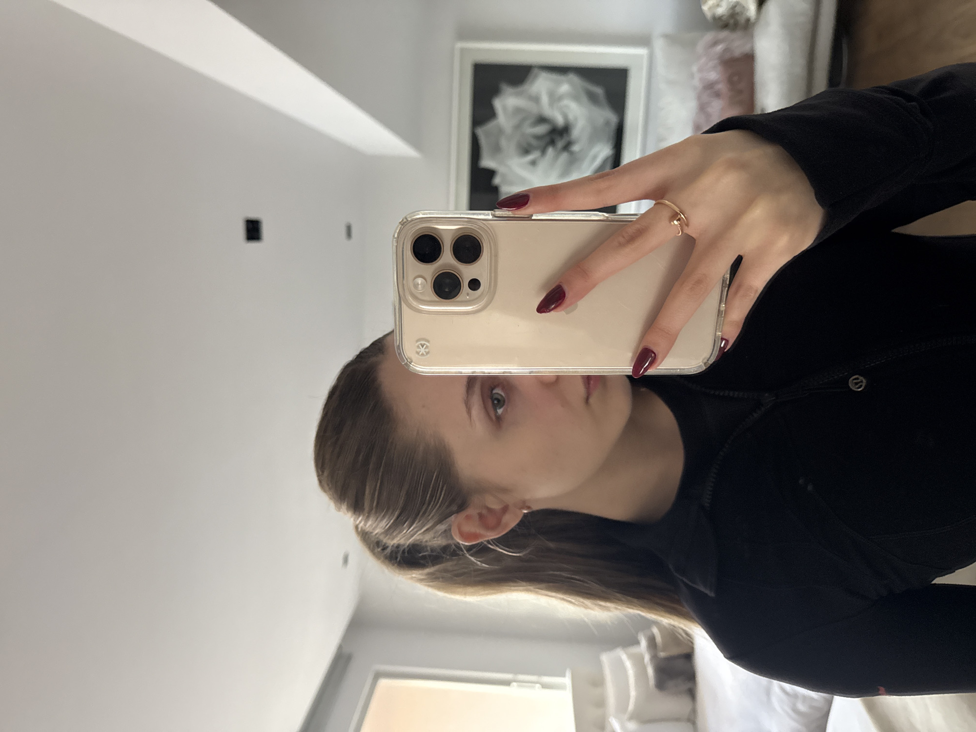 A woman is taking a selfie in what appears to be a modernly decorated room. The walls are painted white. A framed monochrome floral artwork hangs on the wall behind her. She is wearing a black jacket, and she has a ring on her finger. She is holding a smartphone in a clear case. The fingernails are painted with dark red nail polish. The location is unknown, though the prompt suggests Mexico City, Mexico.