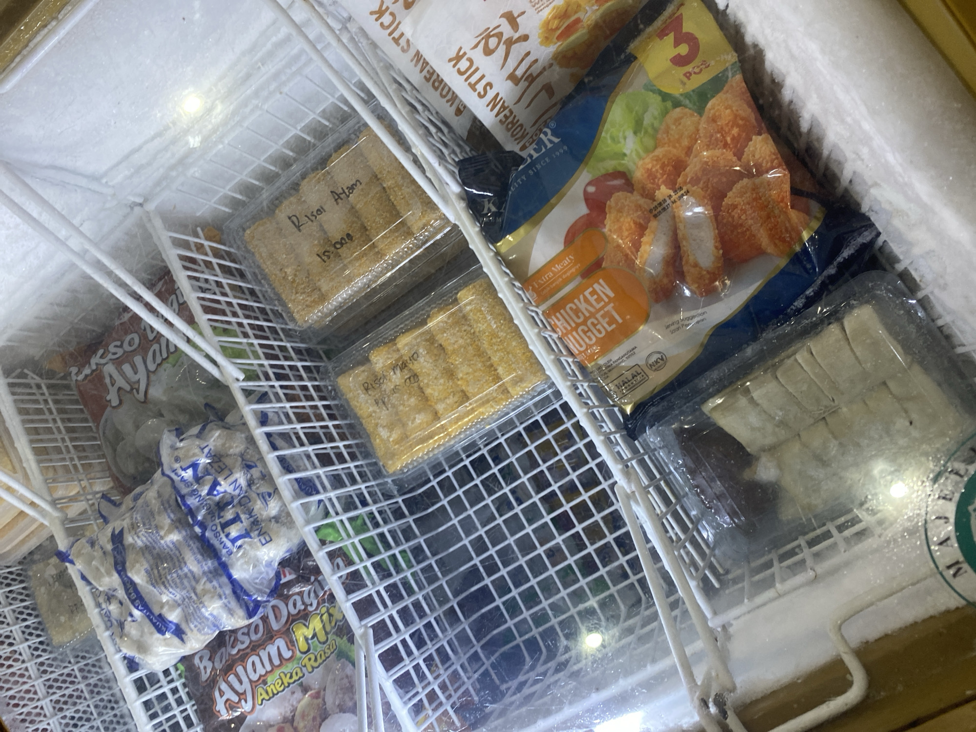 This image shows a close-up, eye-level view of a commercial freezer filled with various frozen food products. The freezer has multiple wire shelves organizing the items. In the top left shelf, a clear plastic bag contains white, round food items, possibly meatballs, with blue text on the packaging. Next to it, another bag with red and brown packaging has a design with green lettering, likely also a meat product.
Moving down, a plastic container holds several oblong, breaded food items. Hand-written text on the lid appears to read "Risol Ayam" and "15.000".
Further to the right and in the next shelf down, a package with a predominantly white and blue design has text that includes "KOREAN STICK". Below this, a package with a bright orange design prominently displays the words "CHICKEN NUGGET" and an image of chicken nuggets with lettuce and tomatoes. The text also indicates "3 PCS".
At the bottom of the frame, a clear plastic container holds multiple rectangular, white food items, possibly dumplings or dim sum. Part of a green circular logo with white text is visible at the bottom of this container, with the visible text appearing to be "MAJE".
The interior of the freezer is covered in frost, indicating its operational state. The lighting appears to be internal to the freezer, with a bright white light source visible in the upper right quadrant. There are no people or any discernible location markers for Payatieng, Indonesia within the image itself.
