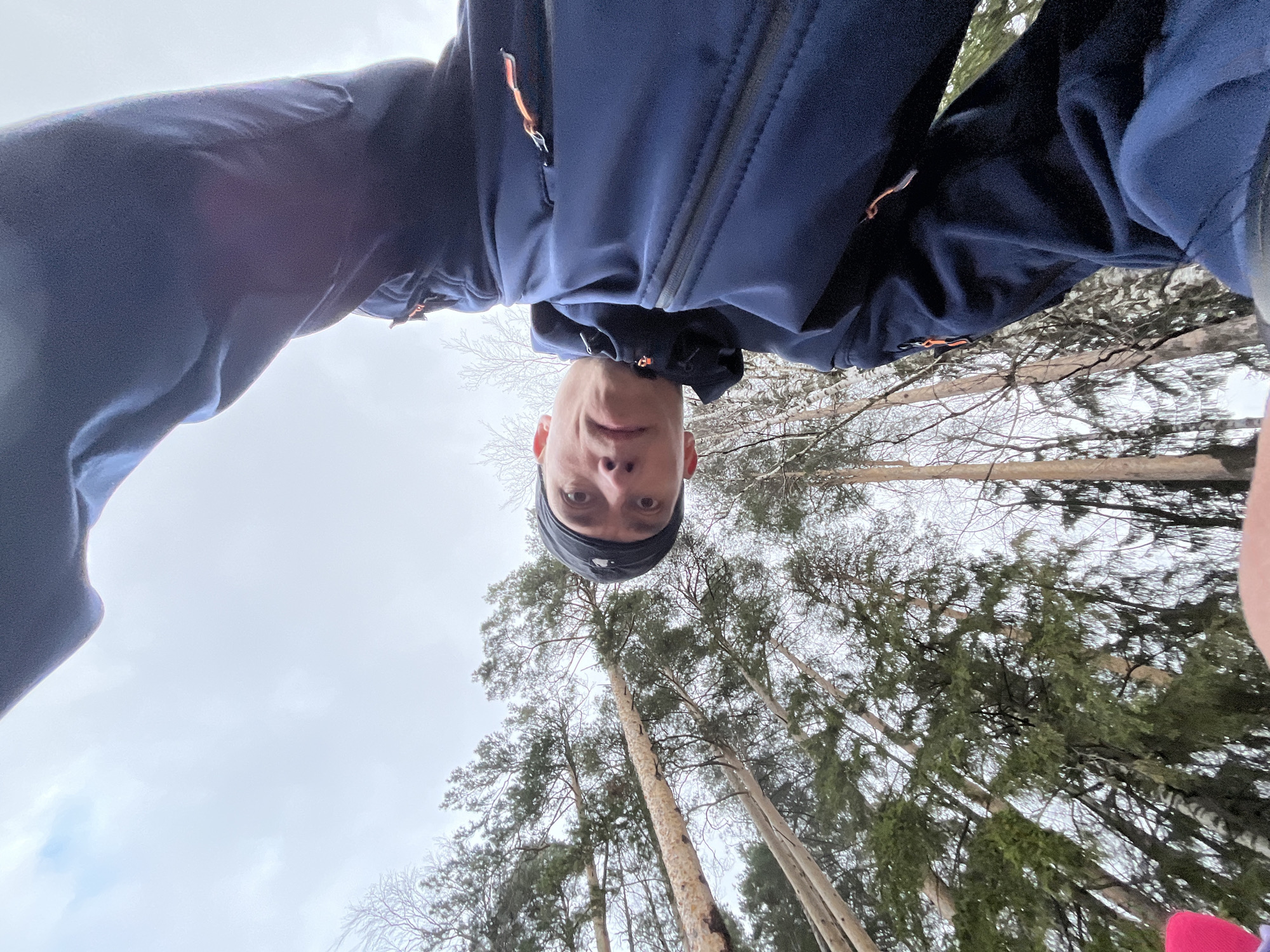 The image presents an unusual low-angle selfie taken outdoors in a forest, likely in Chaikovsky, Russia. The primary subject is a young man, possibly male, wearing a dark blue jacket with orange zipper accents and a black headband or beanie. He is looking directly into the camera with a neutral expression, his face positioned in the lower-middle portion of the frame, viewed from an extreme upward perspective.
The background reveals a natural setting dominated by tall, slender coniferous trees, such as pines, with some bare deciduous branches visible, suggesting a non-summer season. The trees stretch high into a bright, overcast sky, which appears uniformly grey-white, indicating a cloudy daytime. The light is soft and diffused. The unique photographic angle creates a dynamic visual, making the subject appear to be peering down from above, while the tall trees create a sense of vertical expanse. The image captures a moment during an outdoor activity, possibly a walk or hike in the woods. There is no visible text.