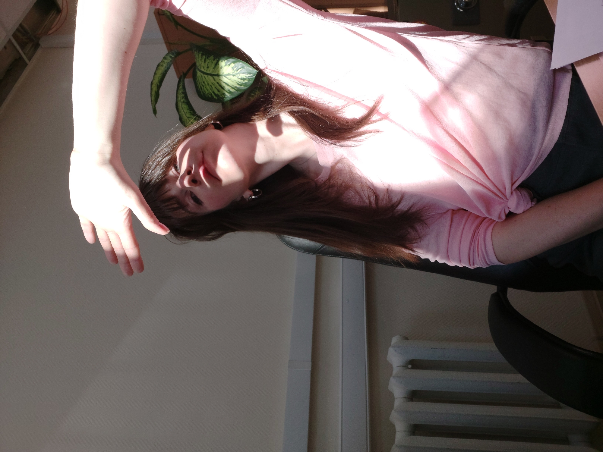 A woman with long brown hair, wearing a light pink shirt and dark bottoms, is seated in a black office chair indoors. Her right arm is raised with her hand extended towards the upper left of the frame. A potted plant with green and yellow variegated leaves is visible behind her. The location appears to be an office setting. The scene is well lit, with sunlight streaming in from the left, creating highlights and shadows. A radiator is visible on the lower right. The location is Chaikovsky, Russia.